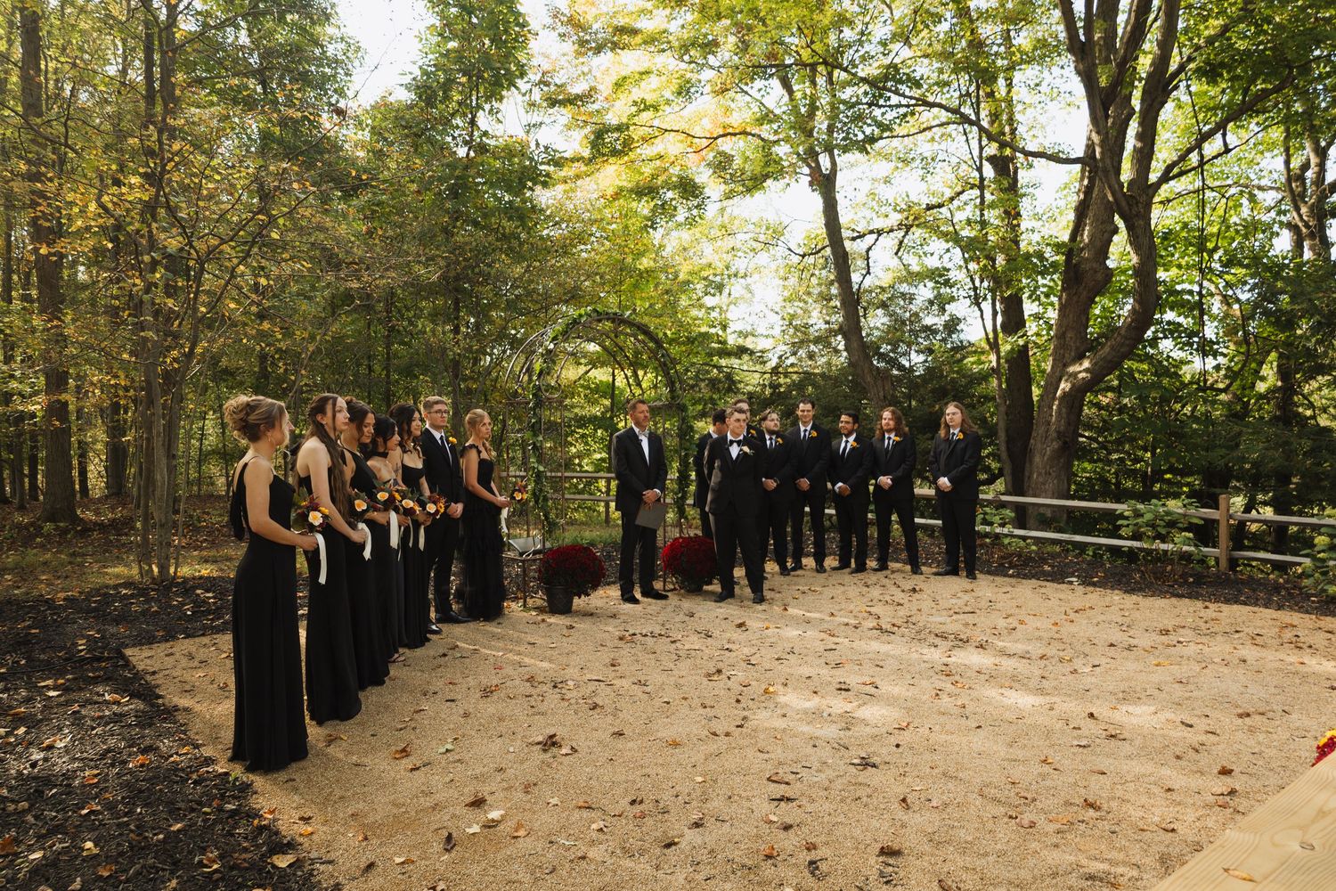 A wedding party in black attire stands in formation during an outdoor forest ceremony.