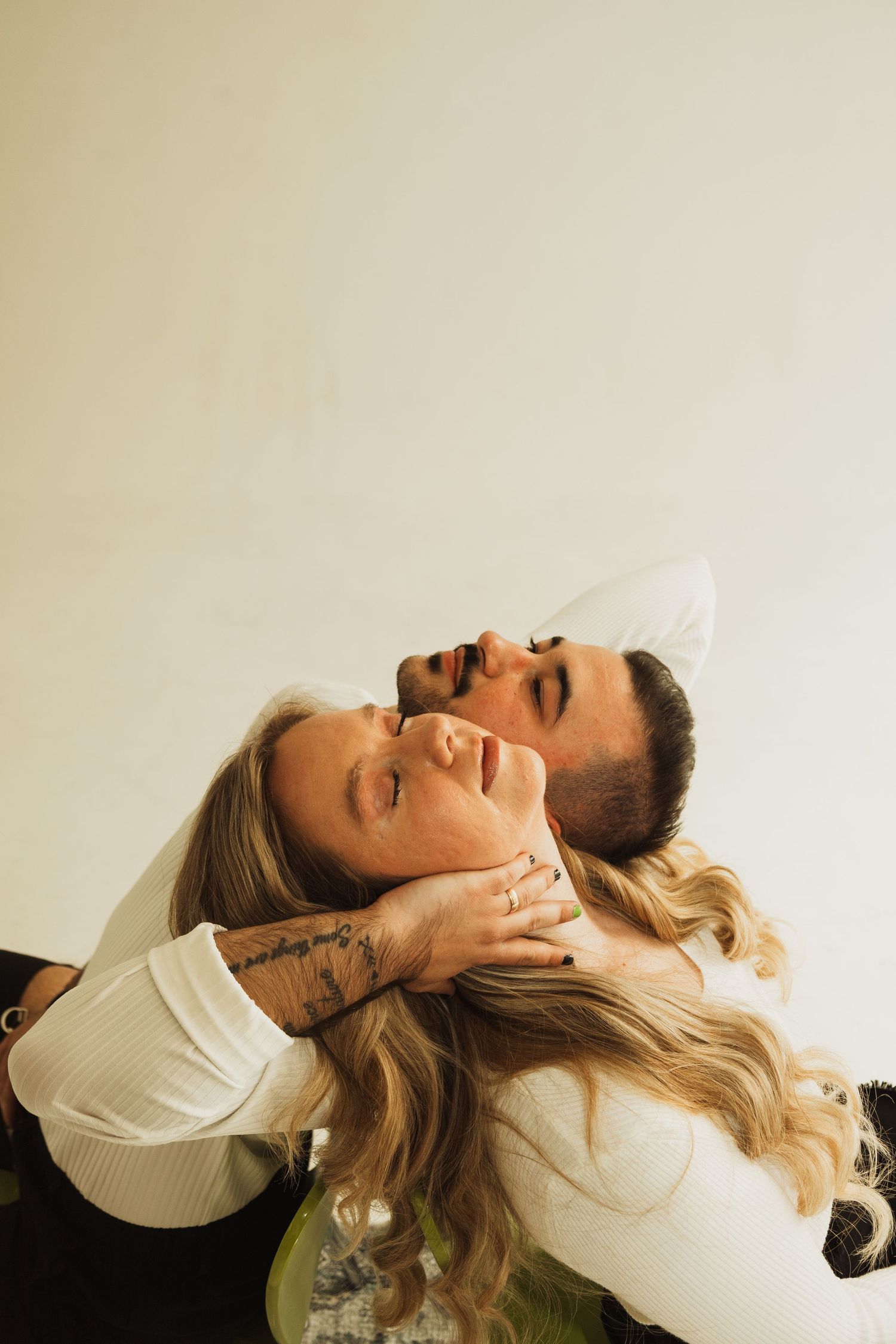 A couple lying on a surface in white tops with long blonde hair and tattoos flashing joyful smiles together.