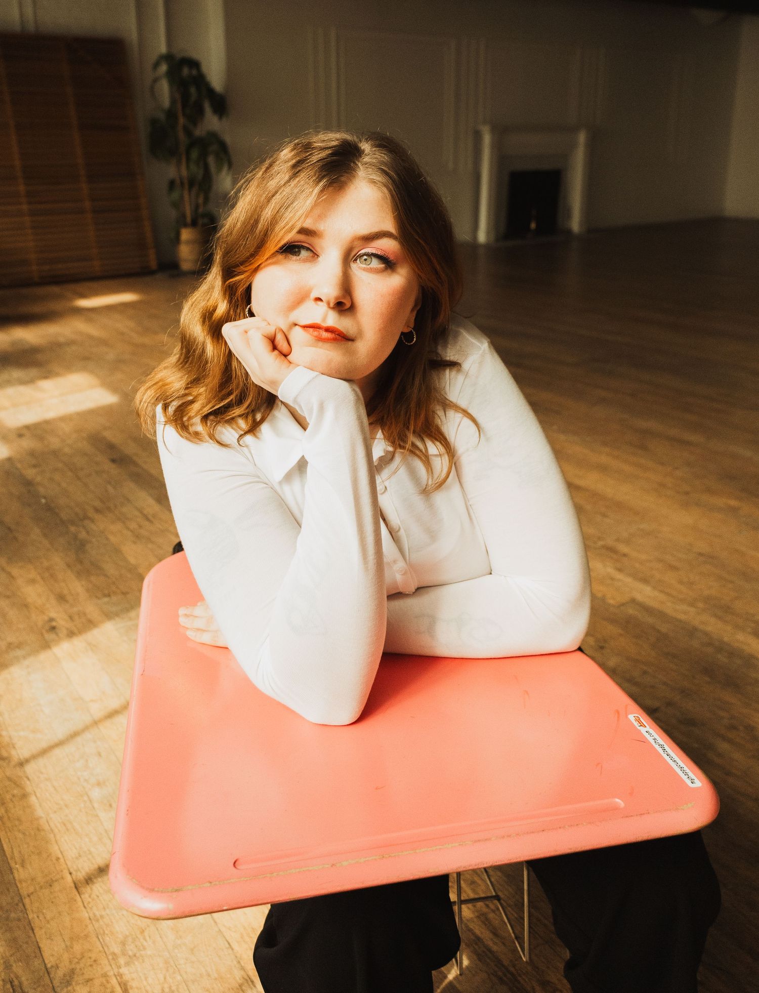 A person in a white sweater leans on a pink chair in a sunlit room with wooden flooring.