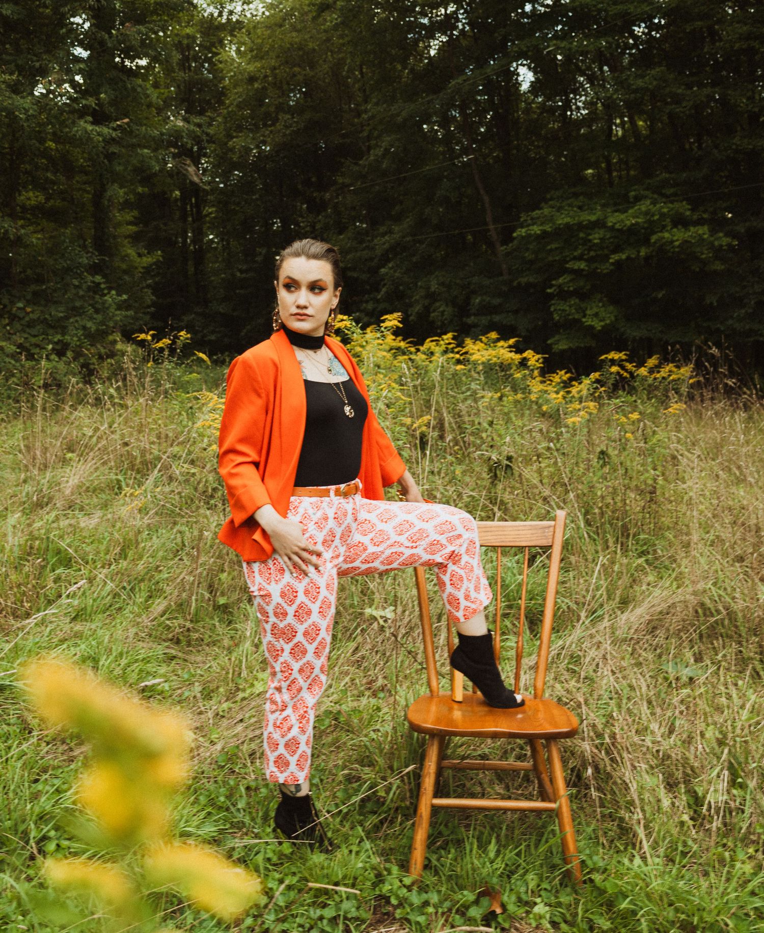 Fashion model poses with wooden chair in field showcasing bold orange and pattern combination.