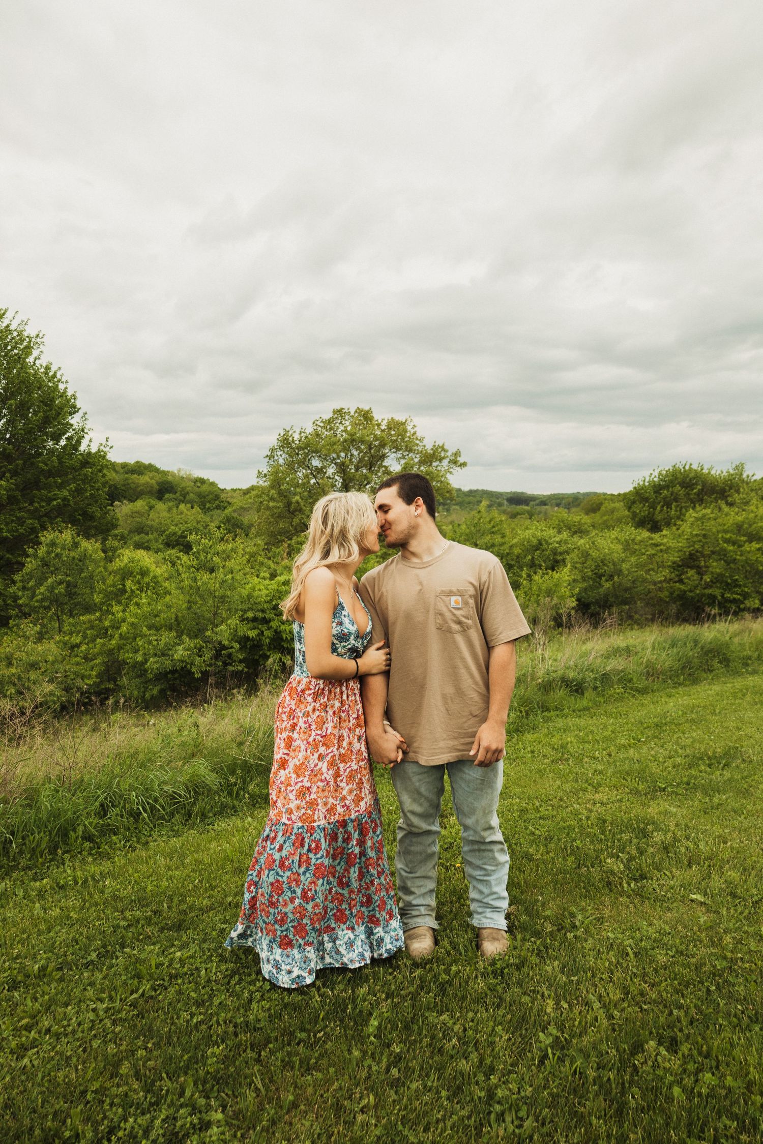 A romantic couple stands in a grassy field on a cloudy day, with her wearing a floral maxi dress and him in casual attire.