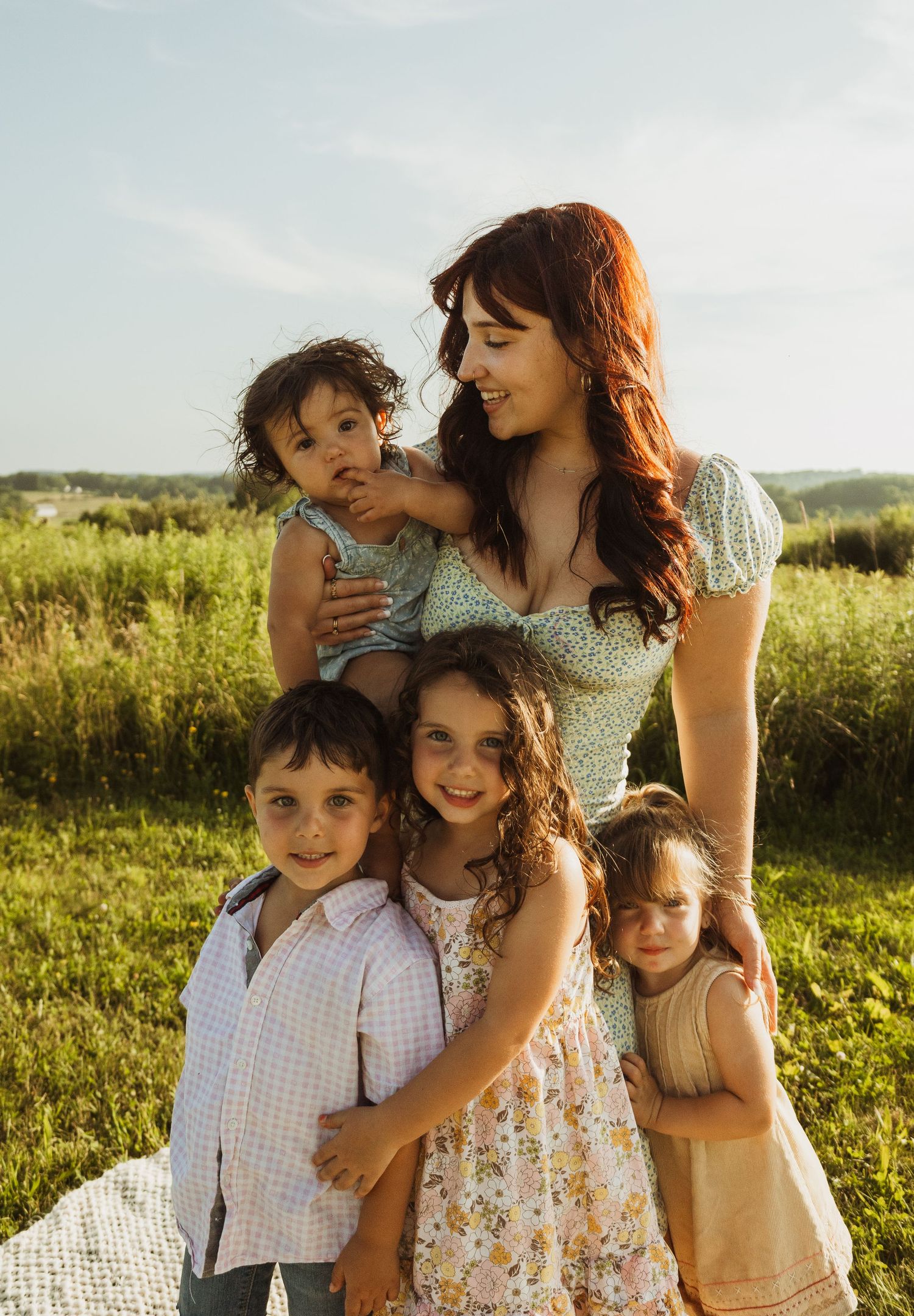 A loving family cuddles together while sitting outdoors in a sunlit grassy field on a warm summer day.