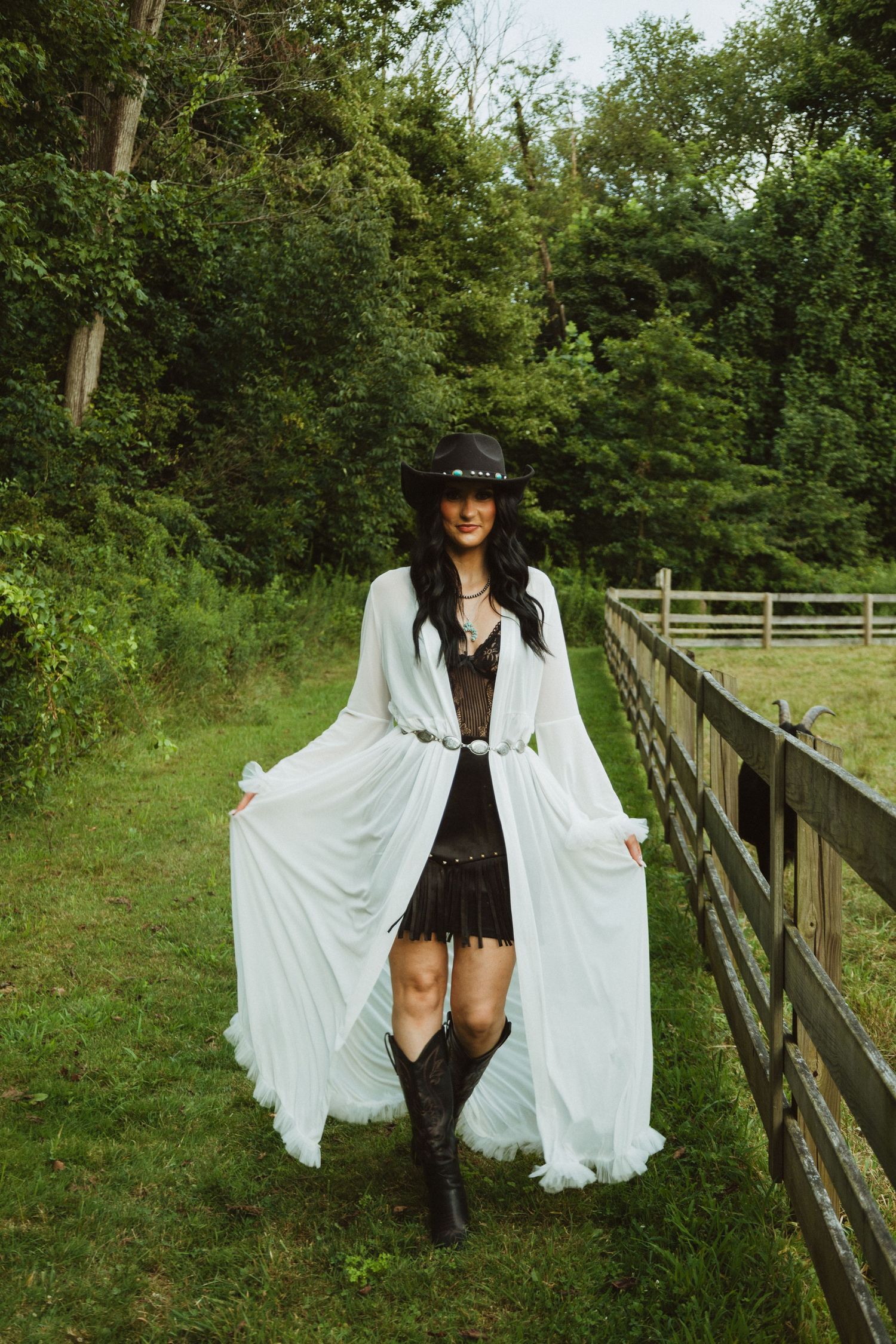A person in a white flowing robe and black hat poses dramatically along a wooden fence in a lush green setting.