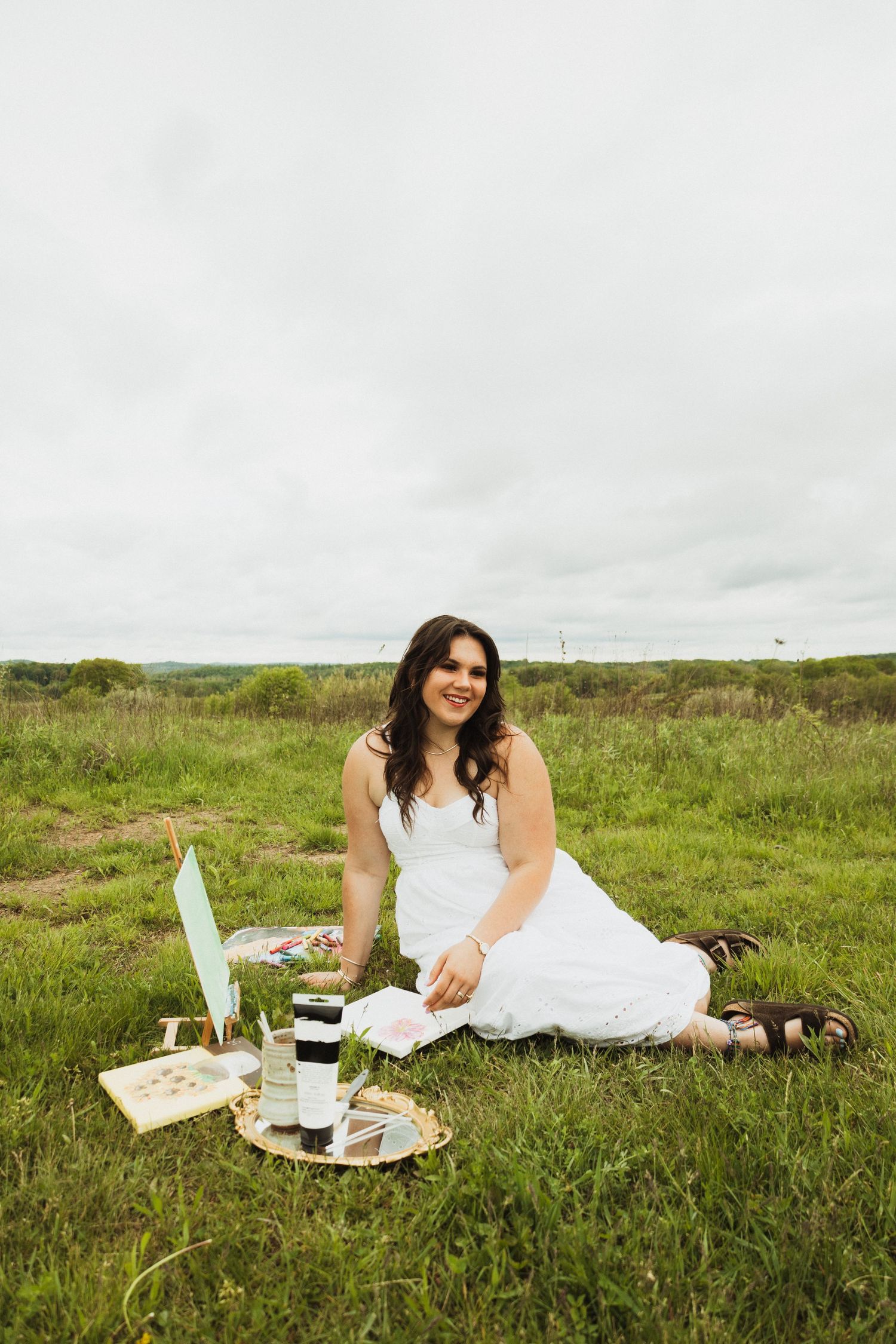 Someone in a white dress enjoys a romantic picnic setup with wine and snacks on grassy hillside under cloudy skies.