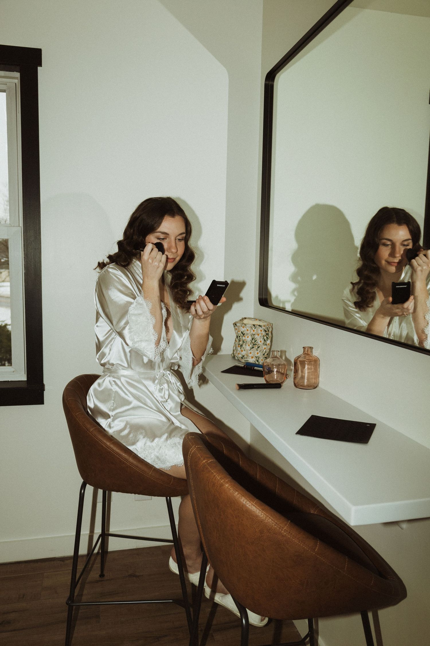 Person in silk robe applies makeup at white vanity with mirror reflection.
