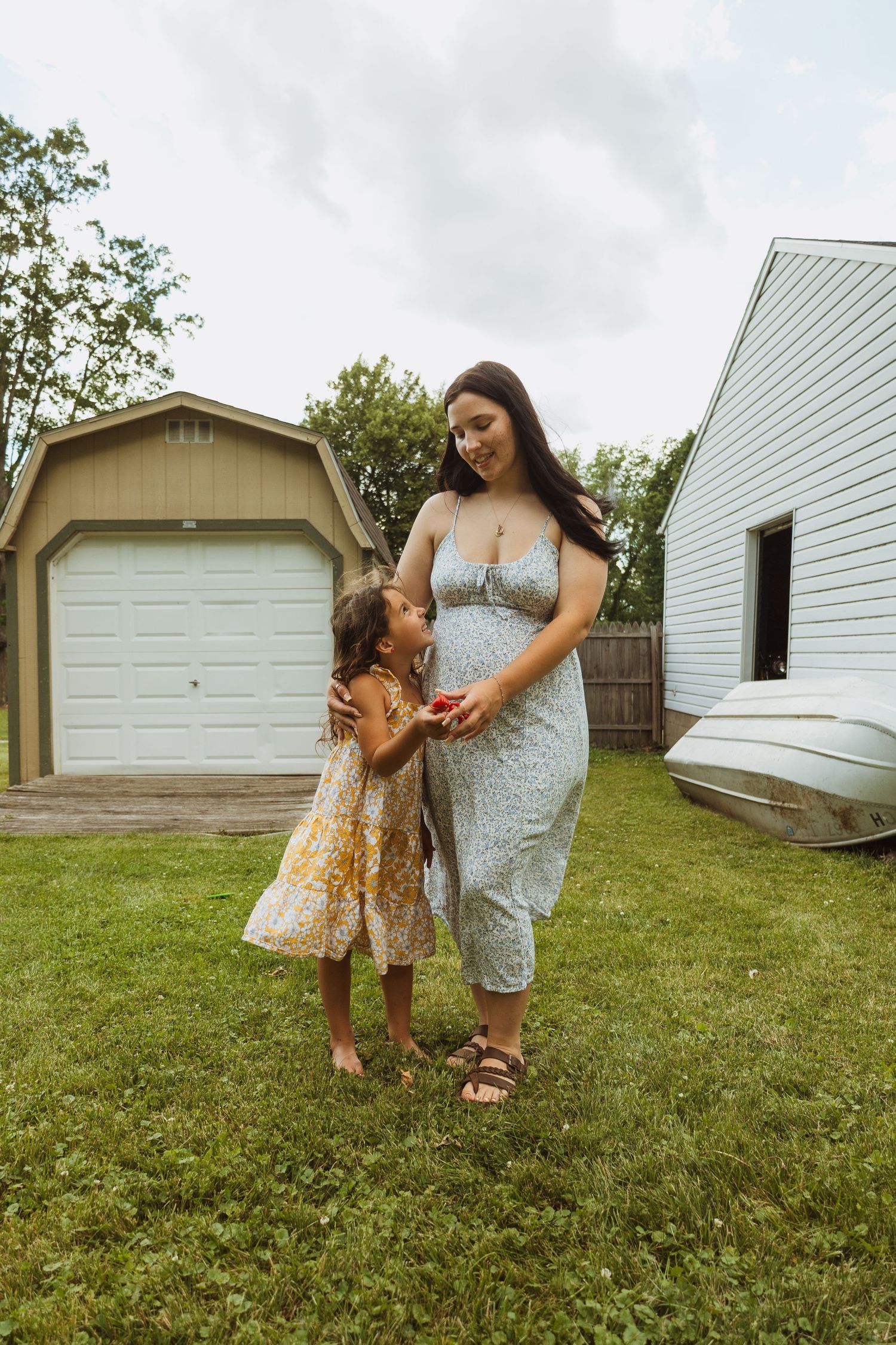 A smiling pregnant woman in a floral dress stands with a young girl in a backyard, sharing a tender moment outdoors.