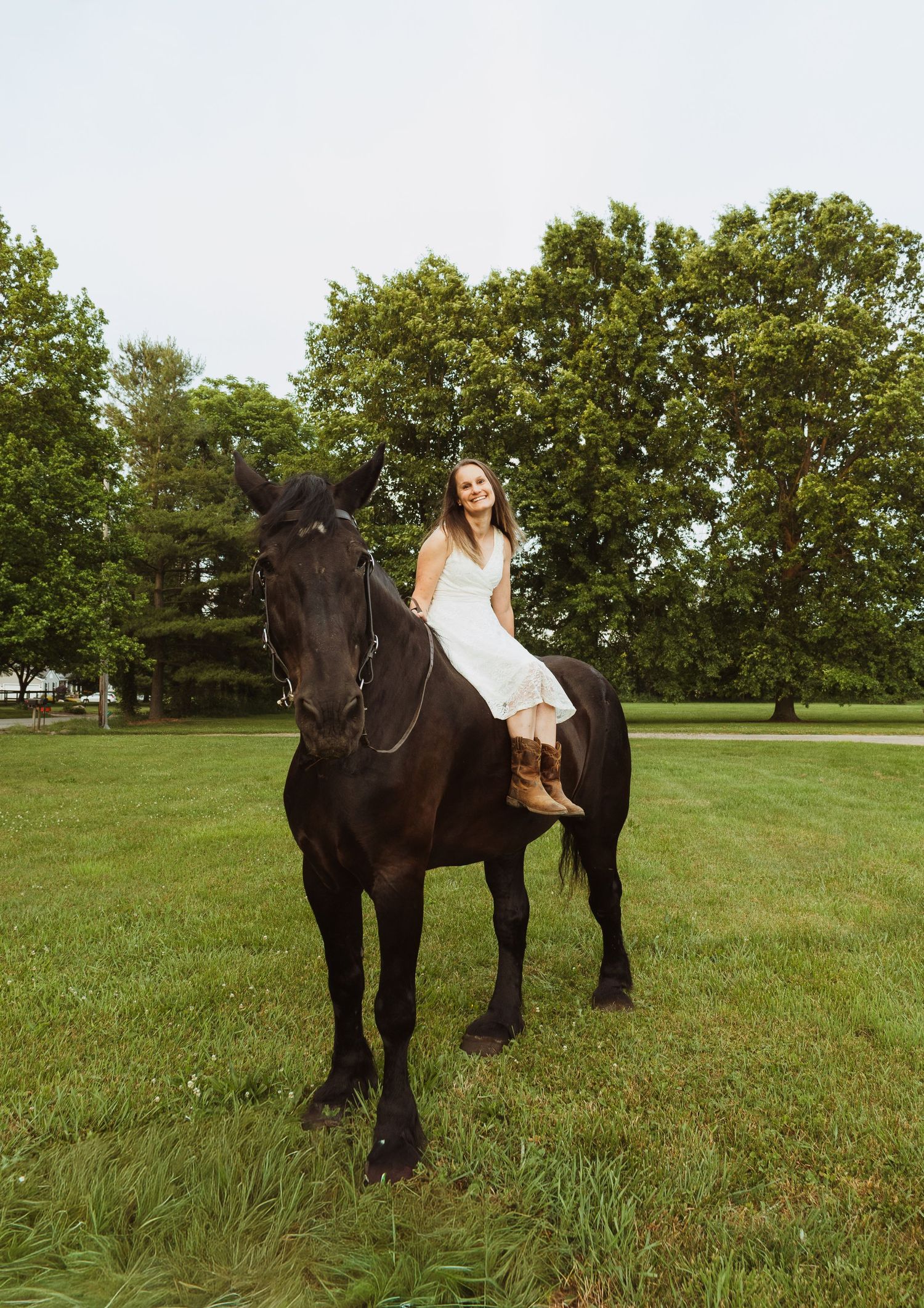 A smiling young woman in a white dress and brown boots rides a black horse on a lush green lawn with trees.