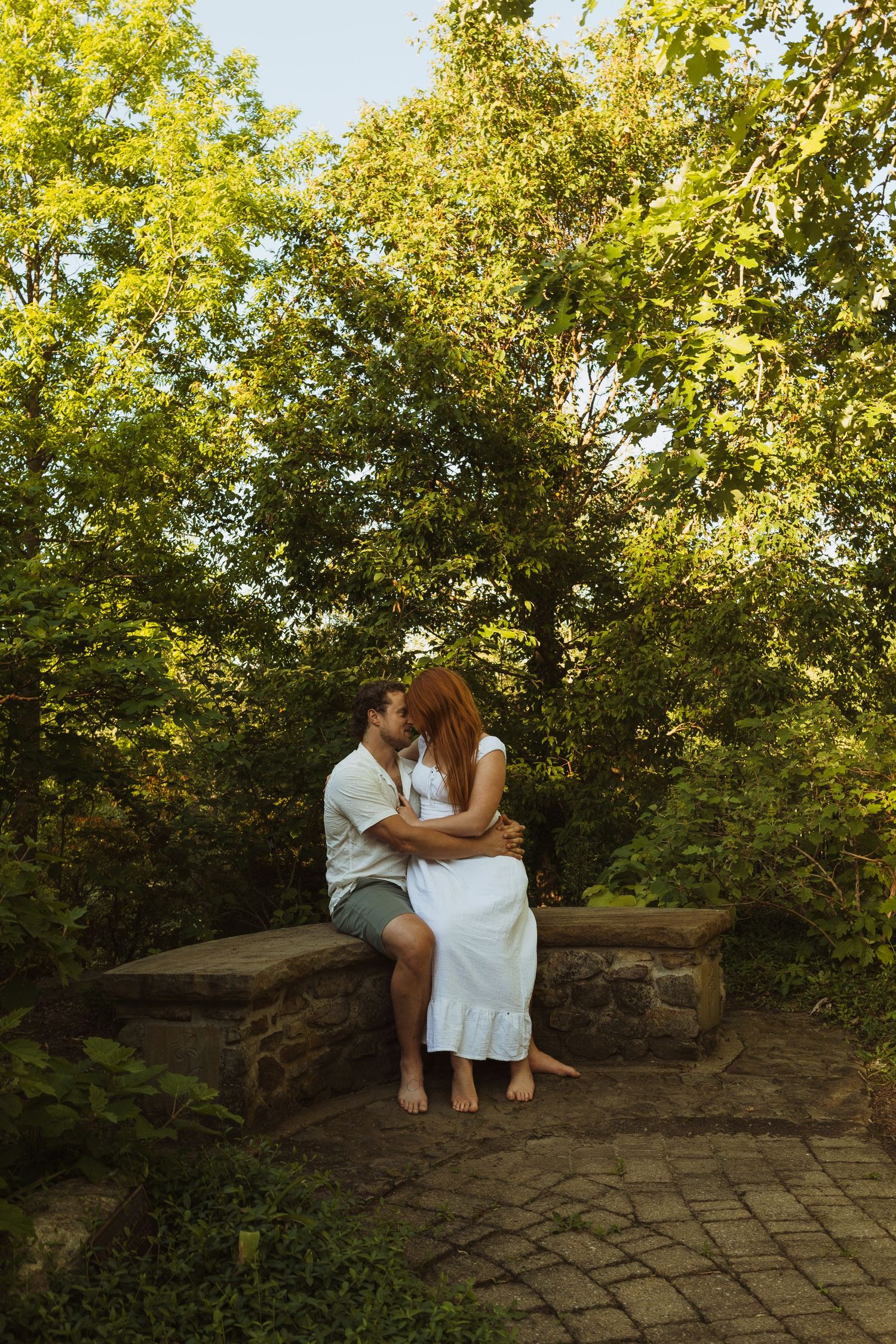 A couple shares a romantic kiss while sitting on a stone bench surrounded by lush green trees in a garden.