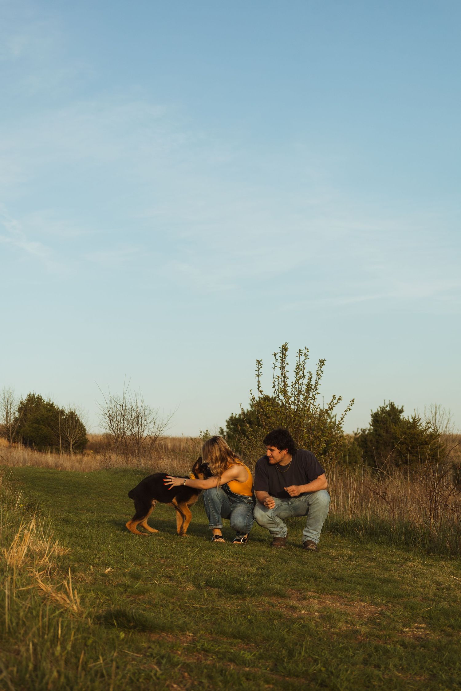 Couple crouching on green grass petting a Rottweiler puppy outdoors with shrubs and blue sky background.