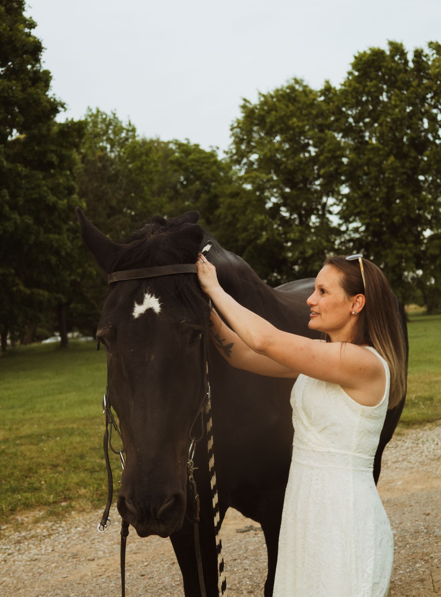 Woman in white dress tenderly grooming a black horse outdoors on a green grassy farm with trees in background.