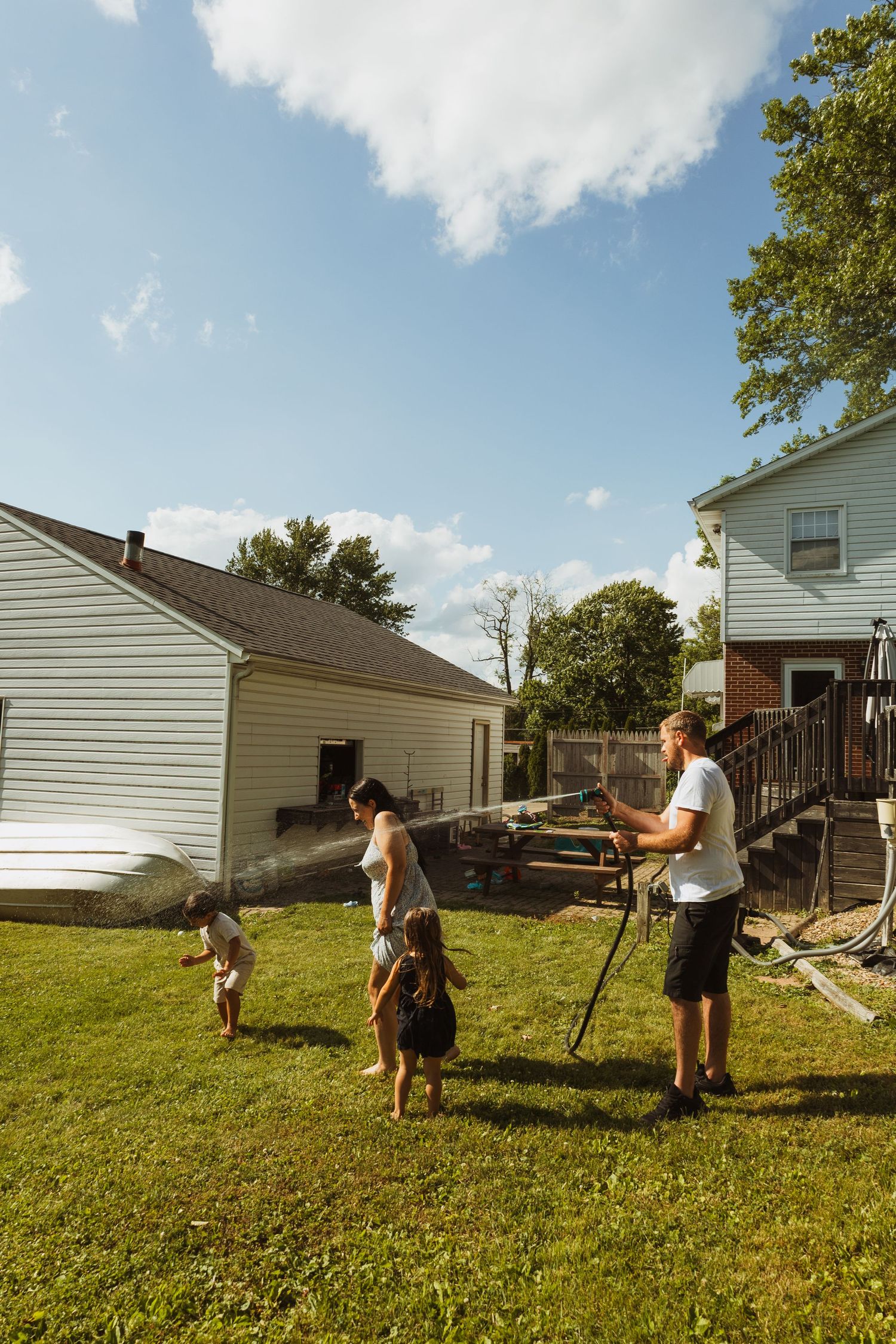 Family playing in backyard with garden hose on sunny day, two houses and green lawn visible in background.