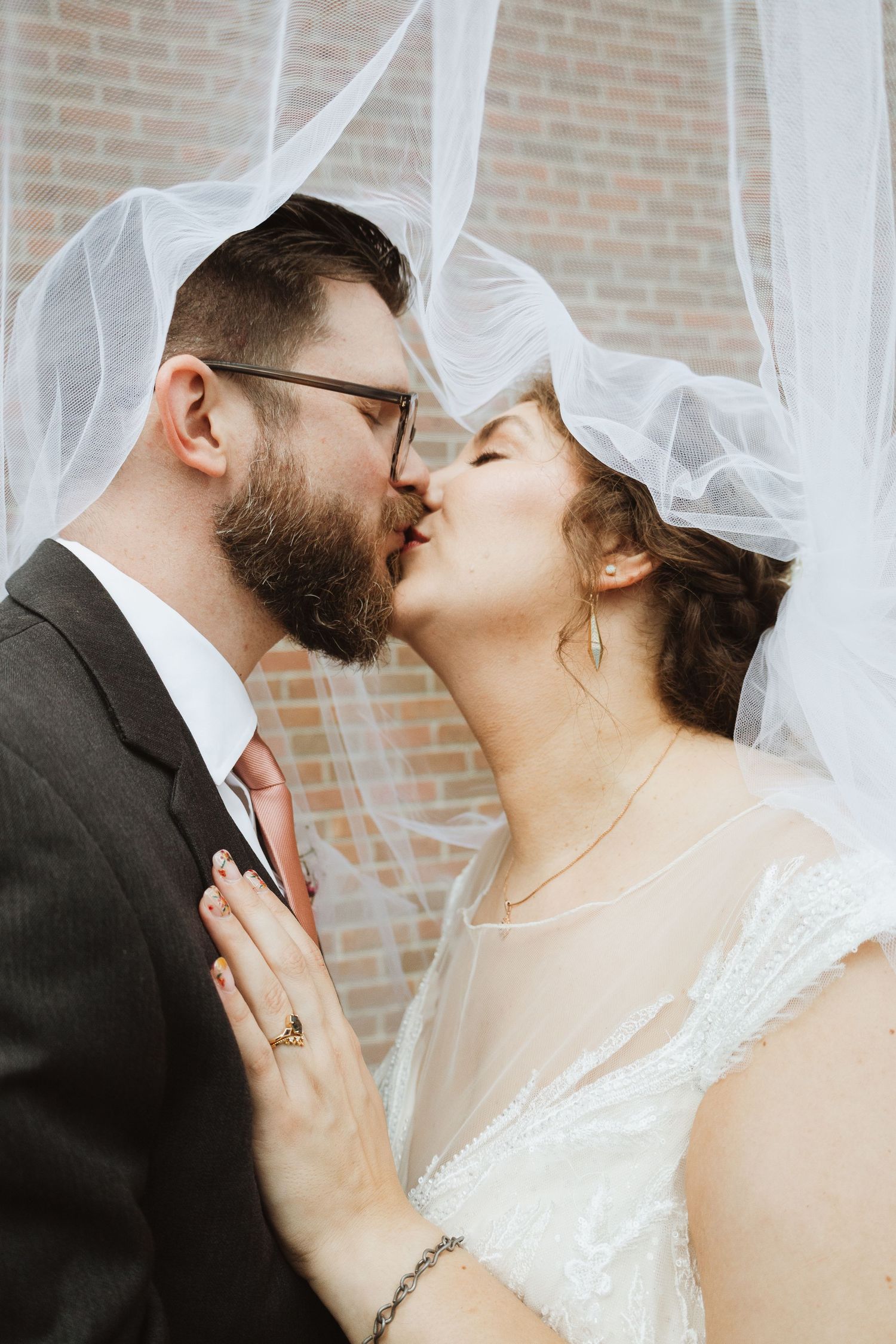 A romantic wedding kiss moment captured against a soft white veil and brick wall backdrop.