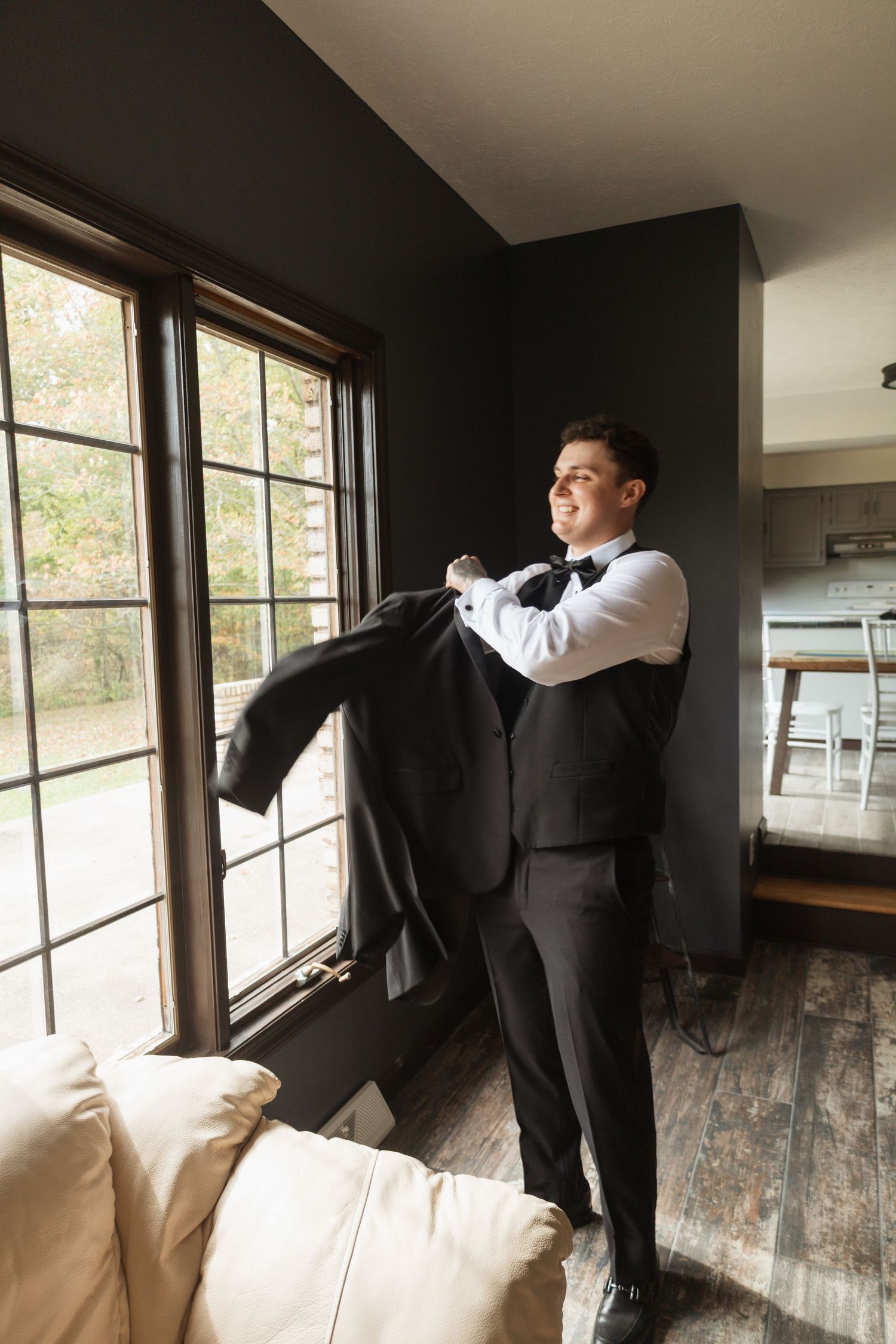 A formal wedding preparation scene with a suit jacket being held up near a window in a rustic room.
