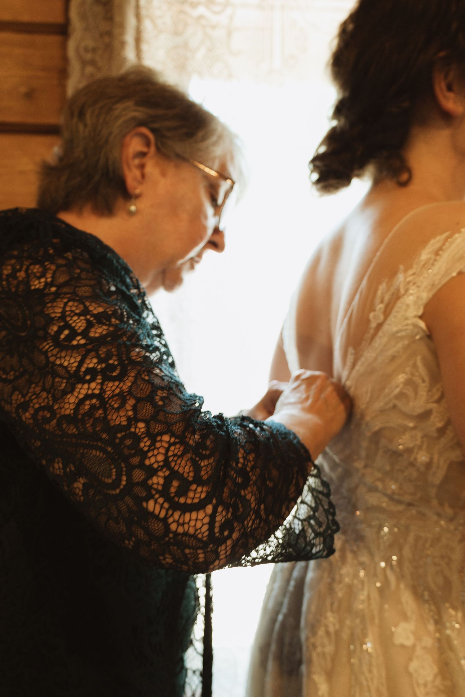 Delicate lace dress being adjusted in soft window light during wedding preparations.