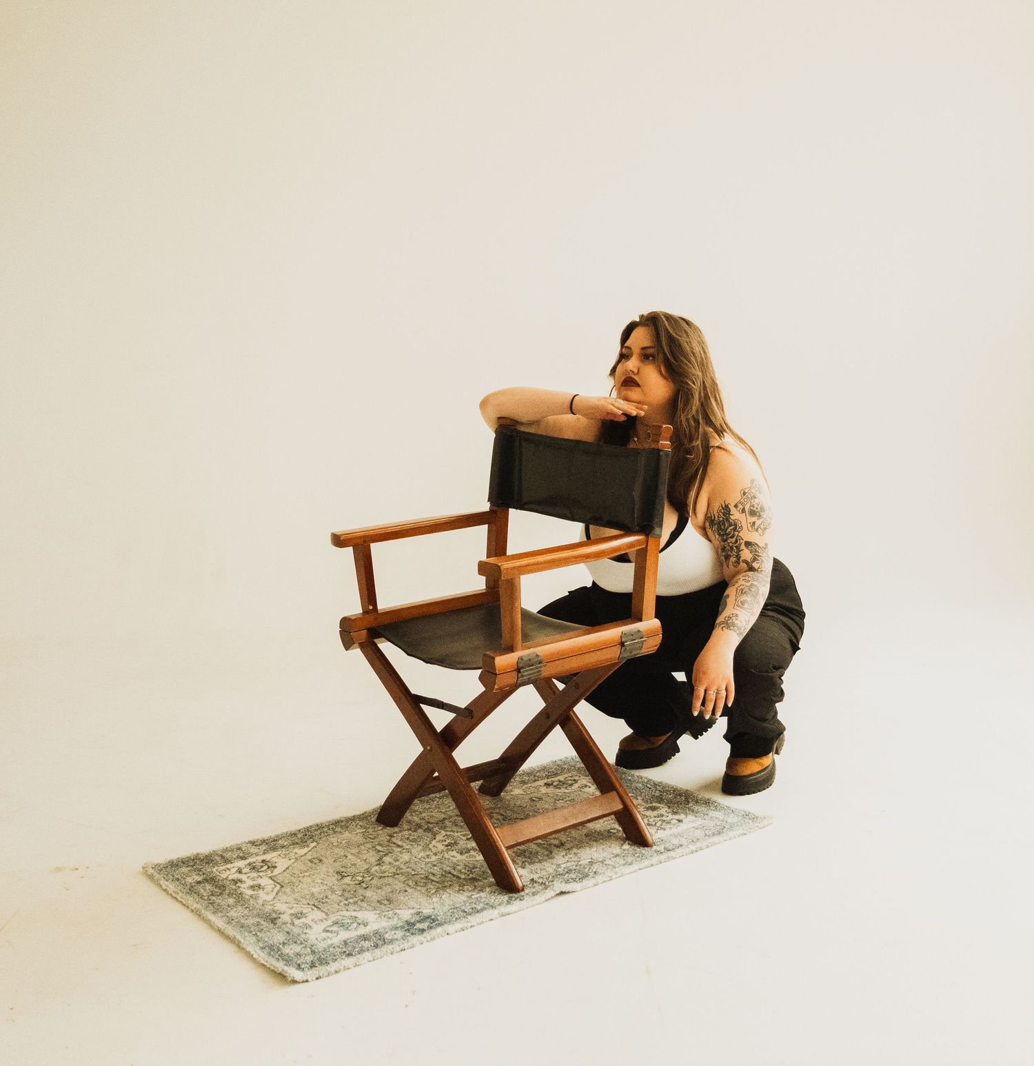 Person crouching next to wooden director's chair on vintage rug in minimalist white studio space.