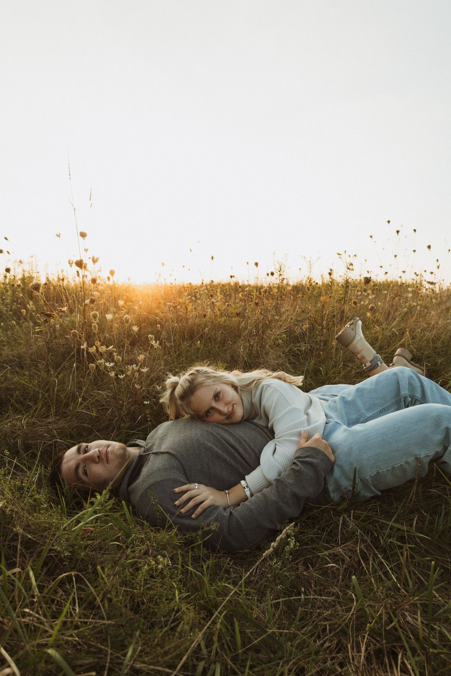 A couple relaxes together in a golden field at sunset with wispy wildflowers overhead.