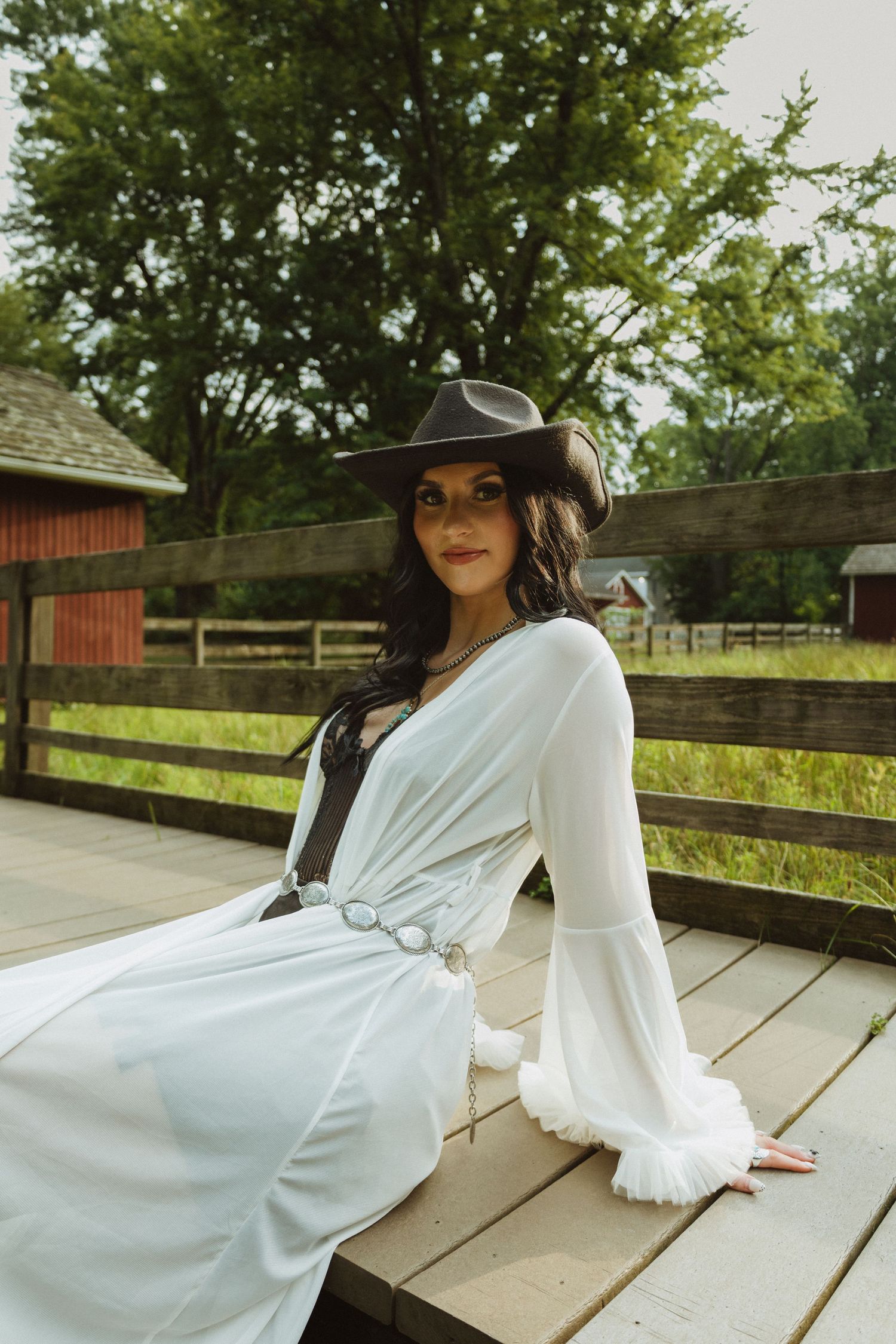 A woman in a flowing white dress and wide-brim black hat poses elegantly on a wooden deck surrounded by greenery.