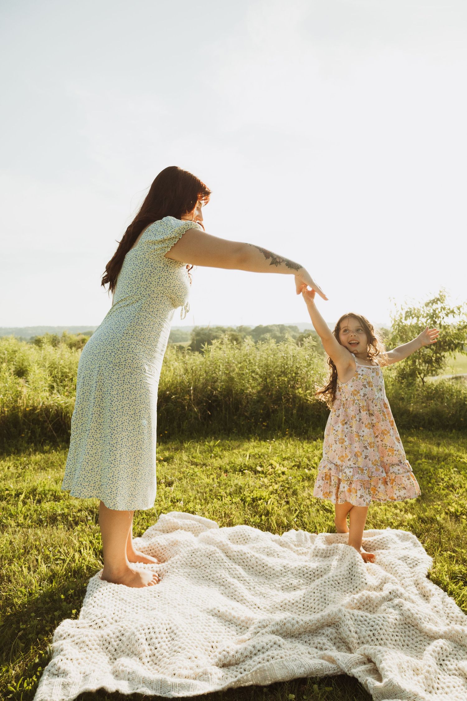 Two people dance and play together on a white lace blanket in a sunny grassy meadow at sunset.