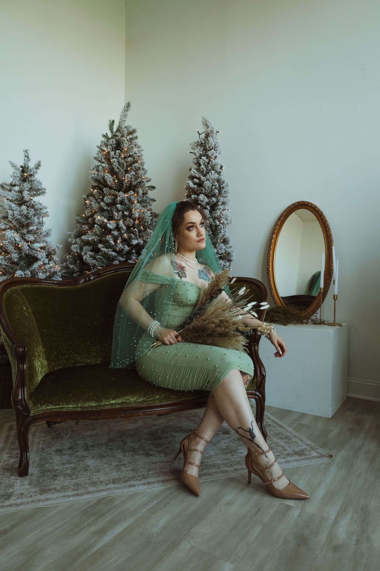 Bride in mint green beaded midi dress and teal veil sits on vintage velvet sofa holding dried bouquet.