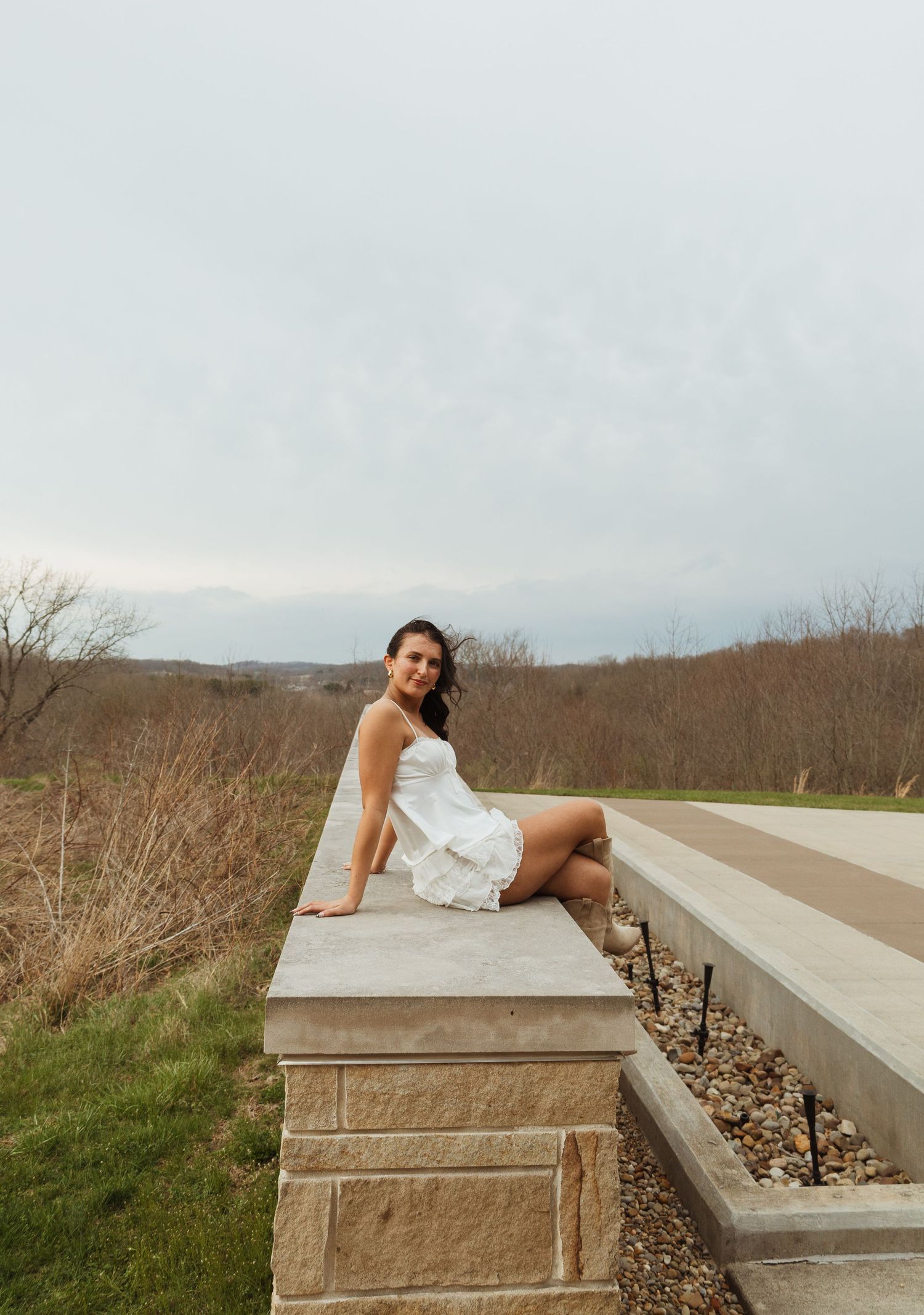 A young woman in a white dress sits gracefully on a stone ledge surrounded by lush greenery under an overcast sky.