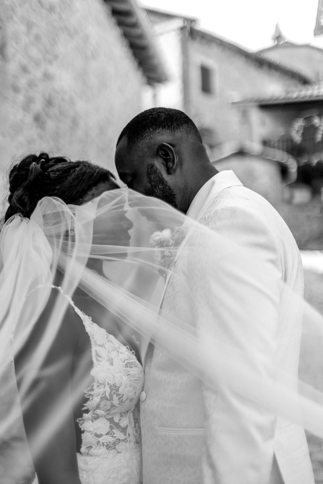 A black and white wedding photograph showing a romantic kiss through a flowing veil.
