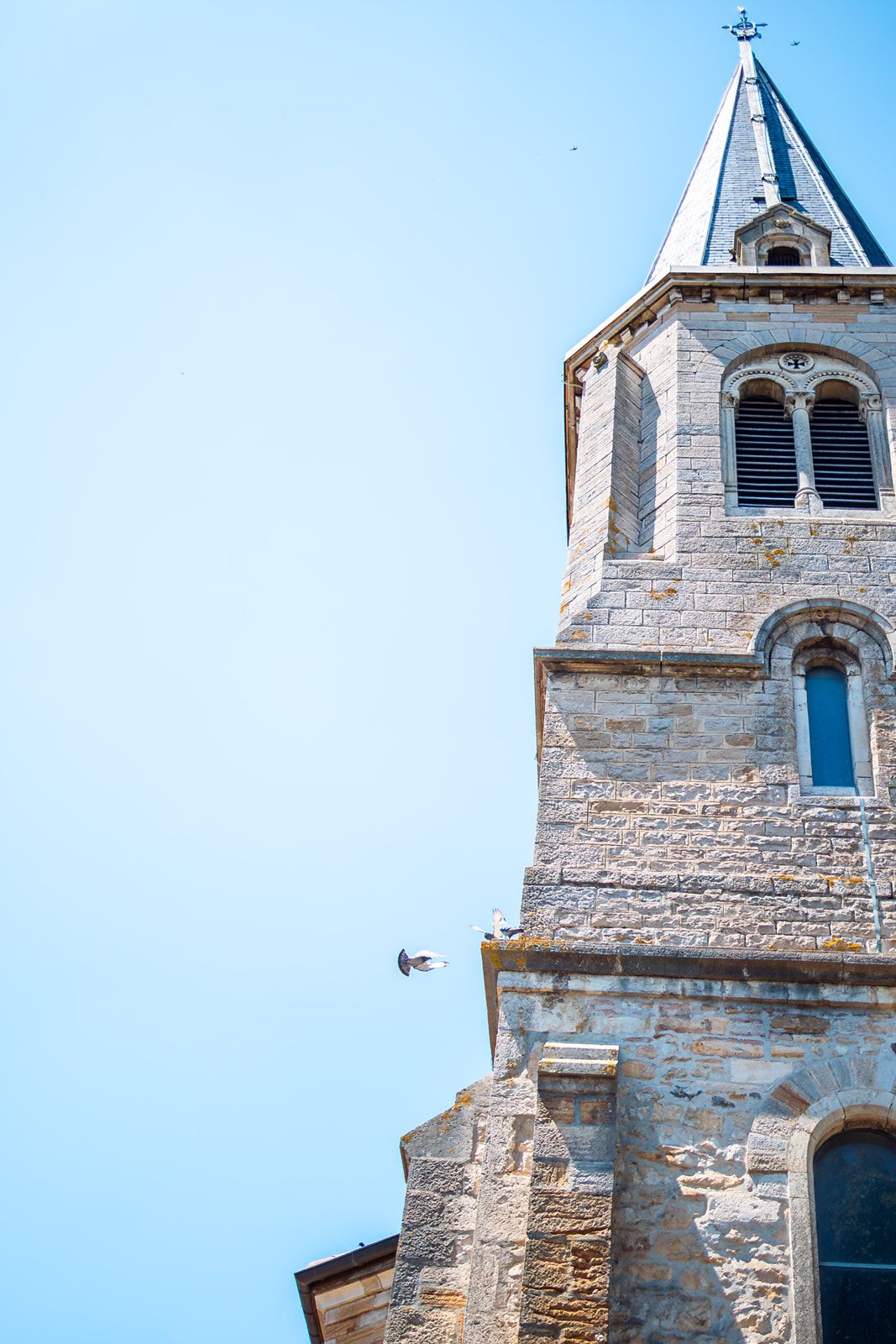 Stone church steeple with cross on top rises against bright blue sky, featuring traditional architectural details.