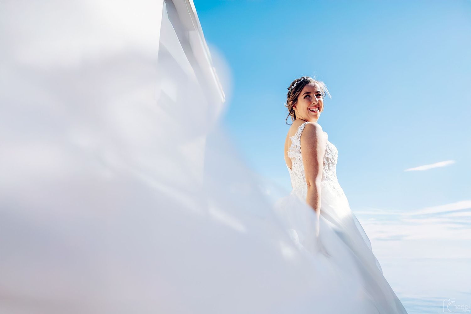 A bride in a flowing white dress stands against a bright blue sky with wispy clouds.