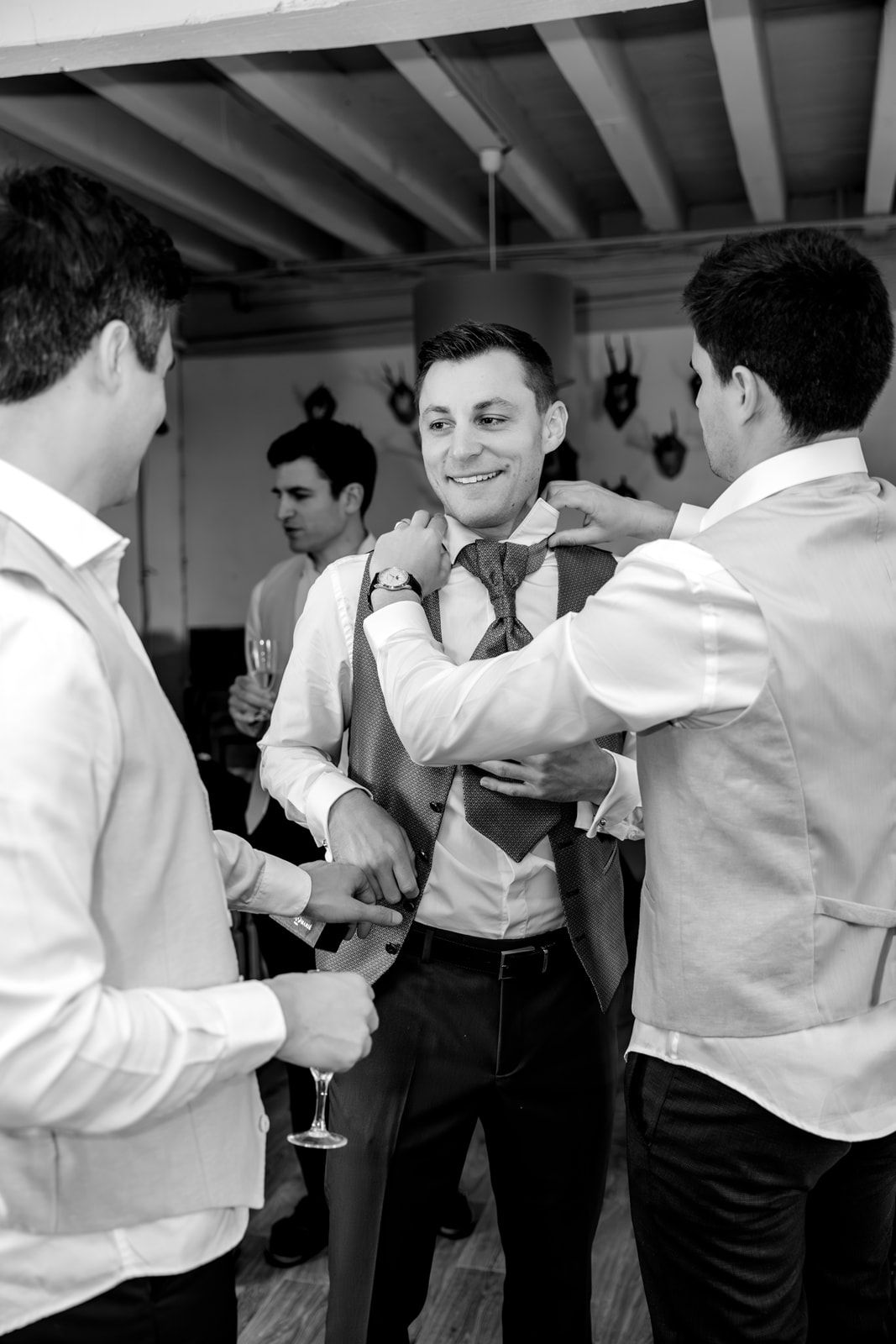 Black and white photo of groomsmen helping groom adjust his tie and vest before a wedding ceremony.