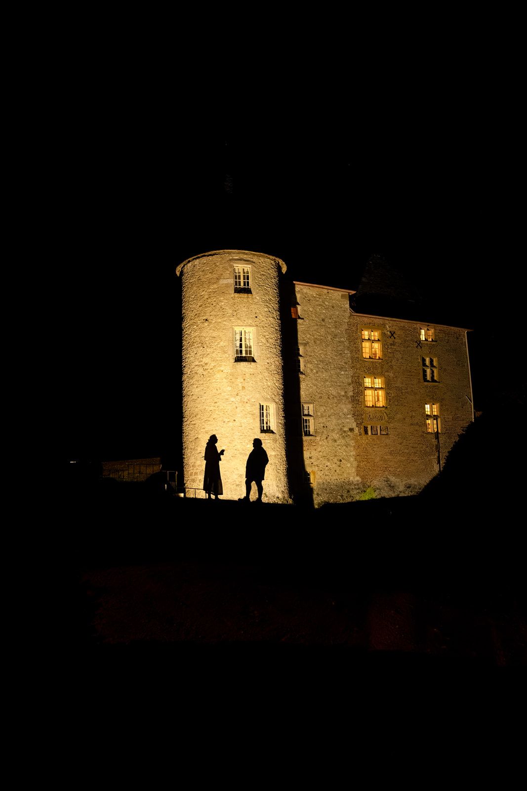 Two silhouetted figures stand before an illuminated medieval stone castle with a round tower at night.