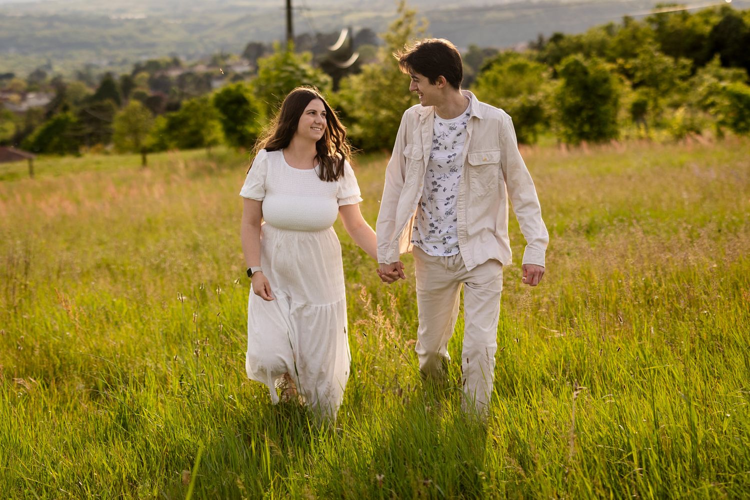 Couple holding hands walking through a sunny green meadow, woman in white dress, man in beige outfit, hills in background.