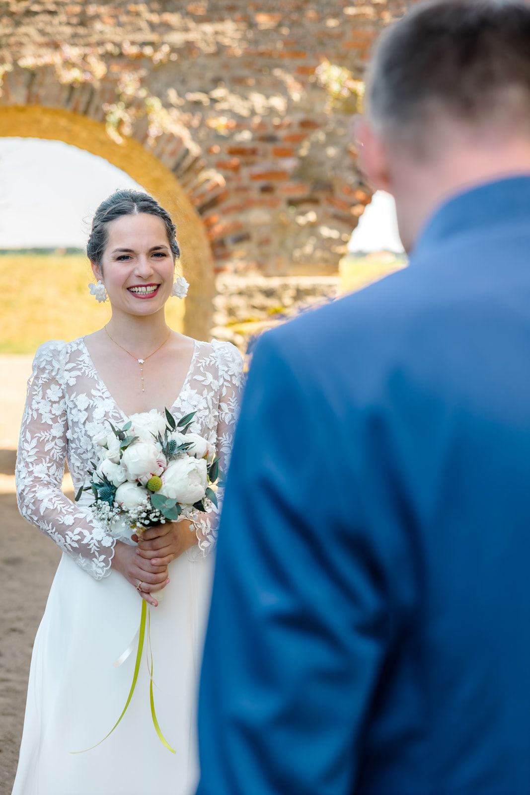 Smiling bride in lace gown holds white bouquet facing groom in blue suit at outdoor wedding ceremony.