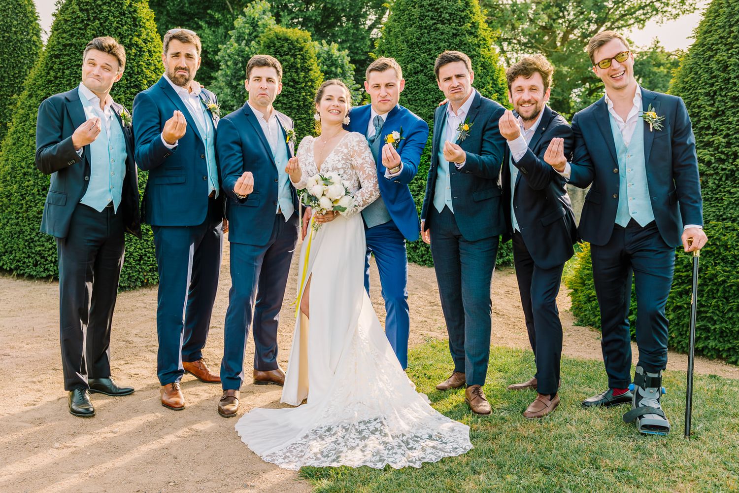 Bride in white lace gown poses with six groomsmen in navy suits, all pointing fingers, in a garden setting.