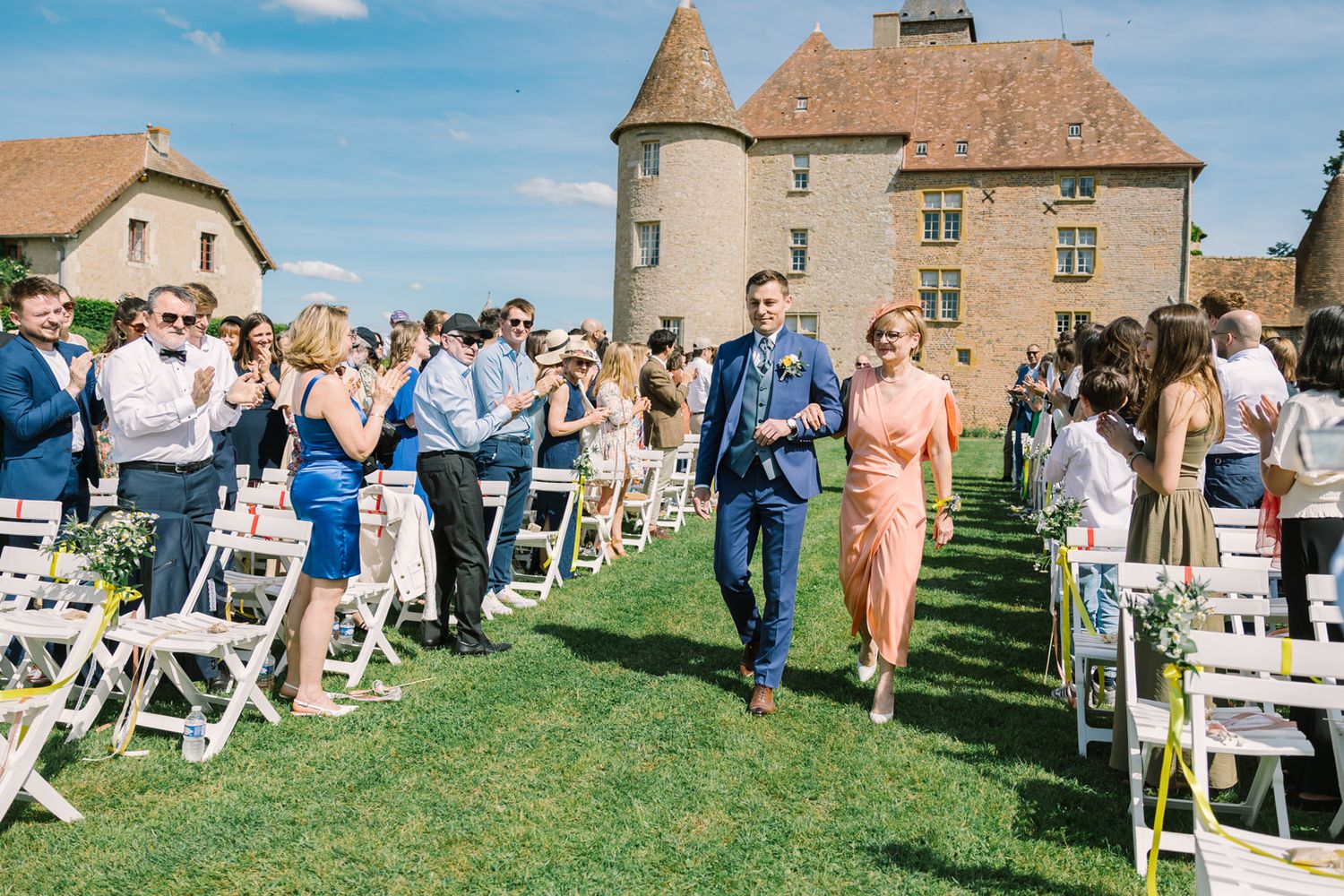 Couple walks down the aisle at an outdoor wedding ceremony on a sunny day with a historic castle in the background.