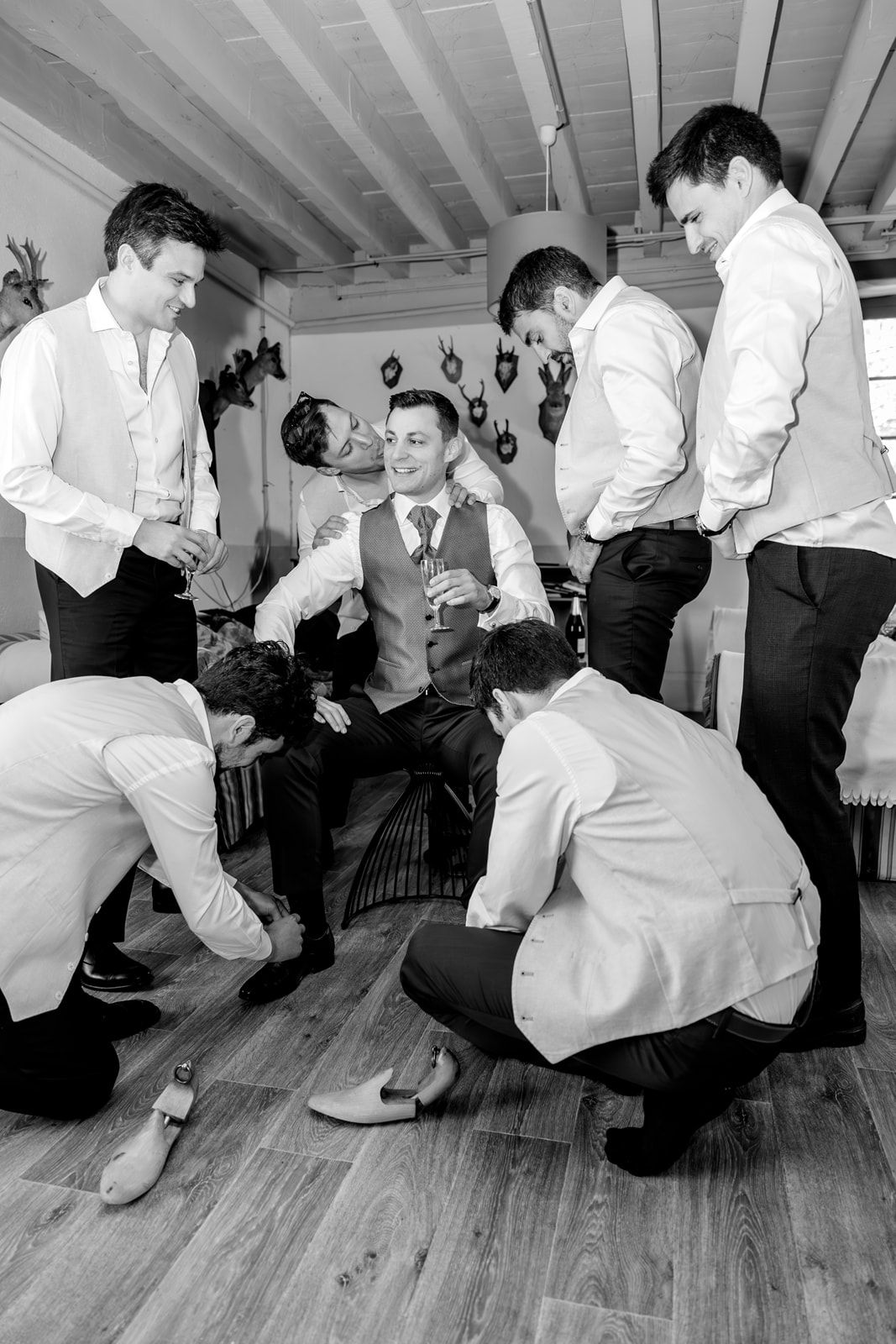 Groomsmen helping groom get ready in black and white photo, adjusting shoes and vest before wedding ceremony.