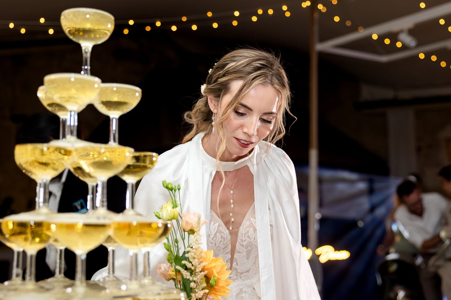 A champagne tower with glasses stacked in tiers glows warmly under string lights at an elegant wedding reception.