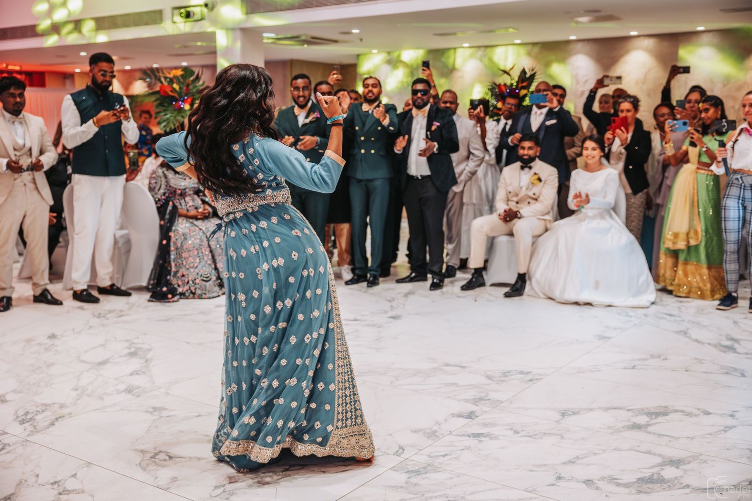 Wedding guests dance in traditional outfits on a white dance floor during reception celebrations.