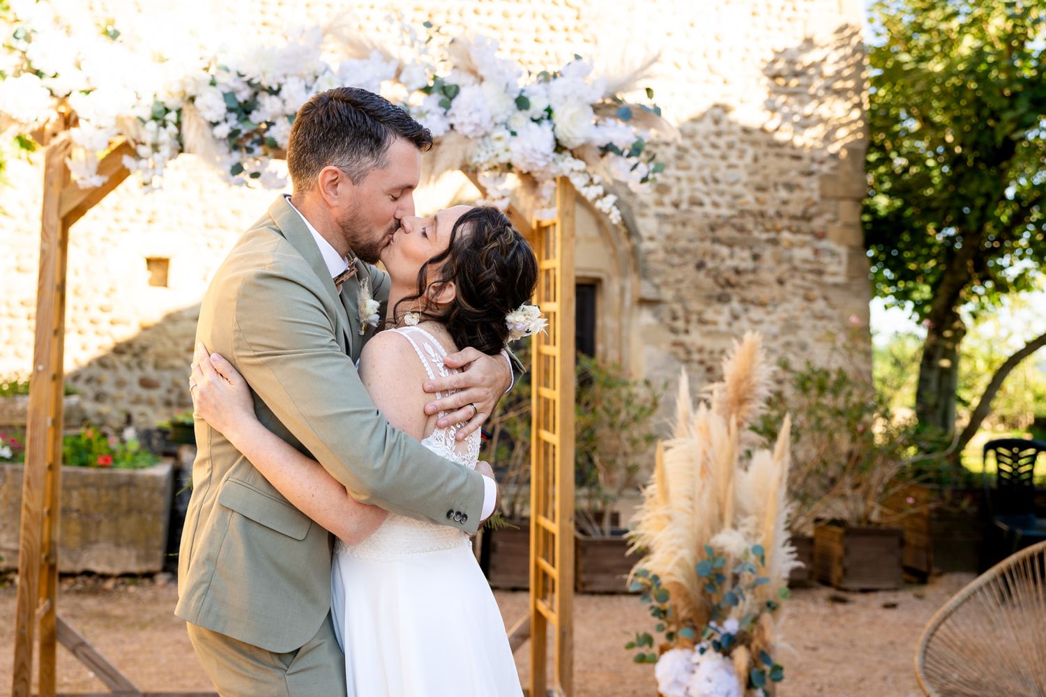 A romantic wedding couple embraces under a floral arch at a rustic stone venue during golden hour.