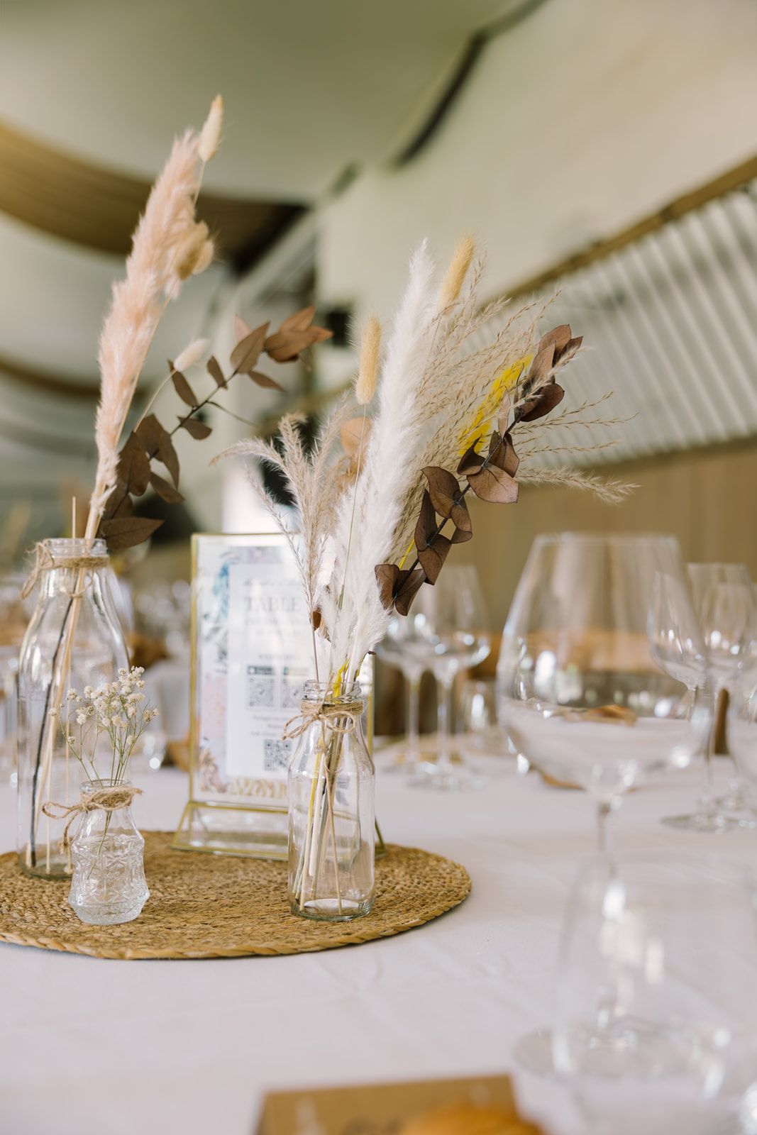 Elegant boho wedding table decor with dried pampas grass in glass bottles on woven placemat with crystal glassware.