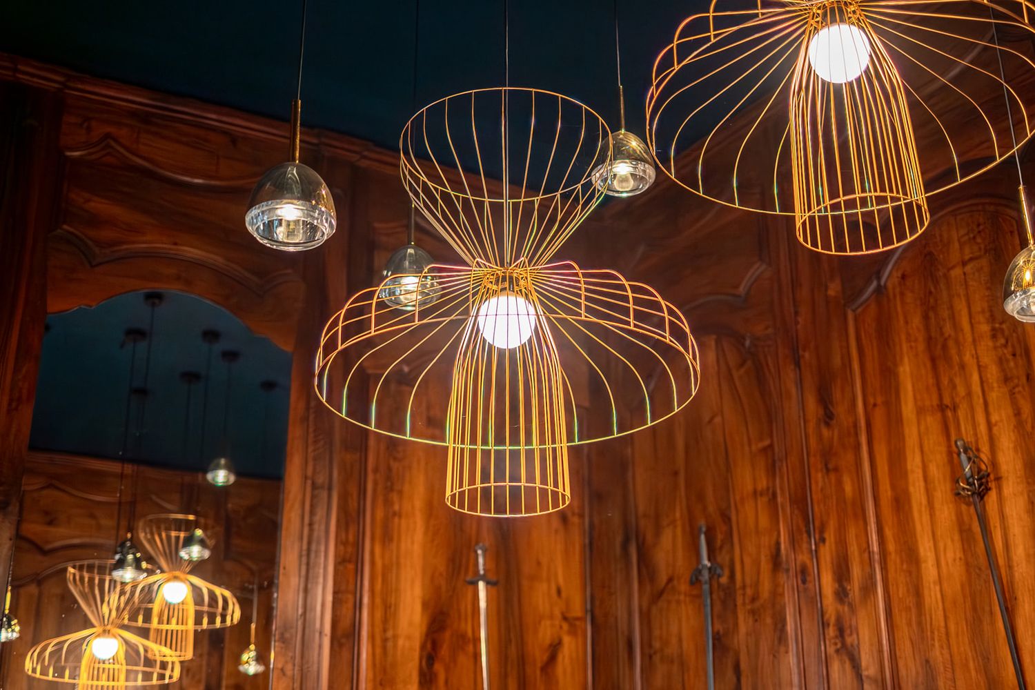Modern wire cage pendant lights with glowing bulbs hang from wooden ceiling in warm ambient interior setting.