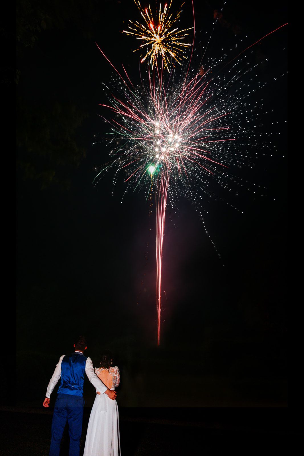 Bride and groom watch colorful fireworks light up the night sky at their wedding celebration.