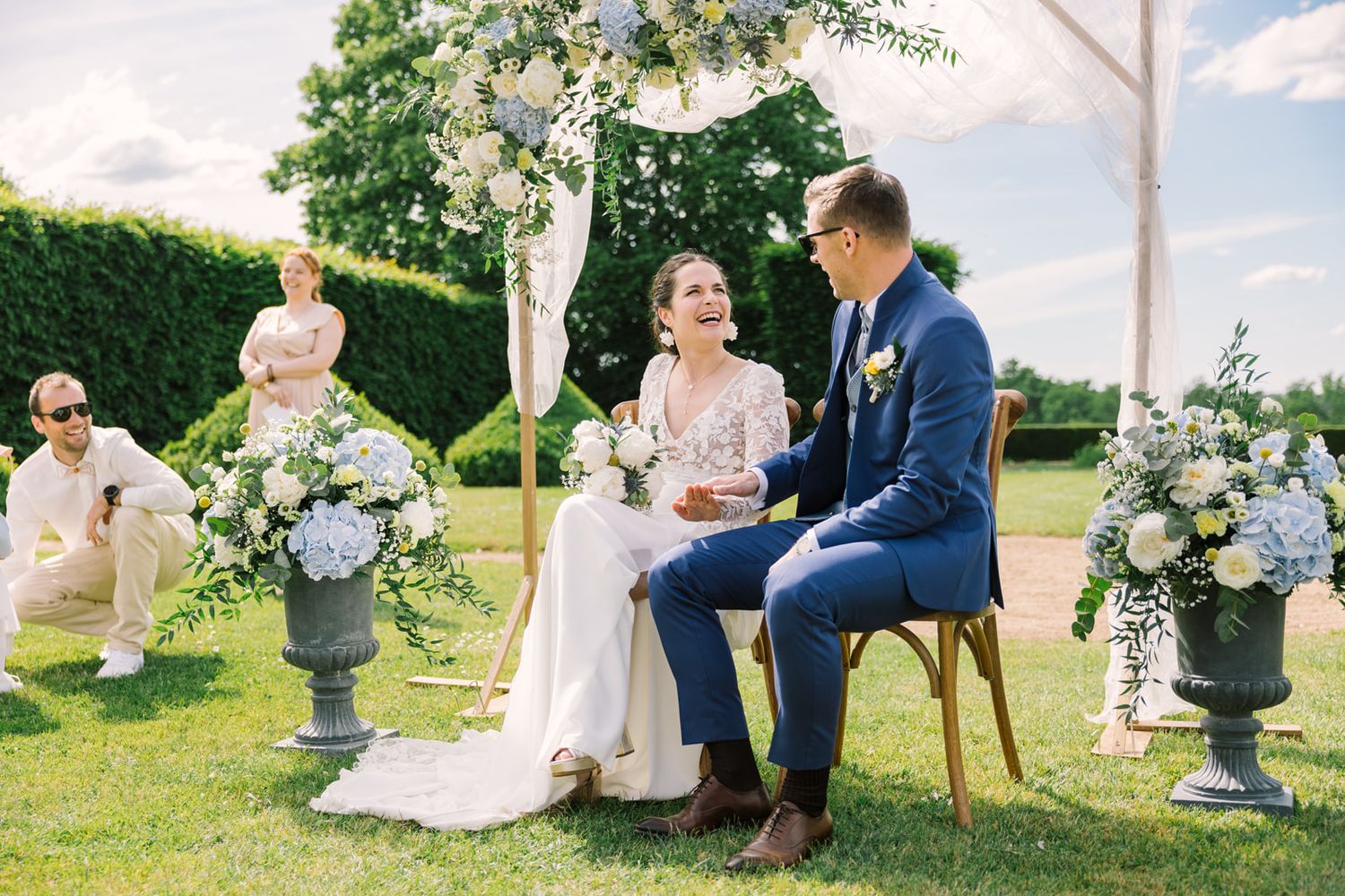 Bride and groom laughing during outdoor wedding ceremony with blue floral arrangements and lush green garden backdrop.