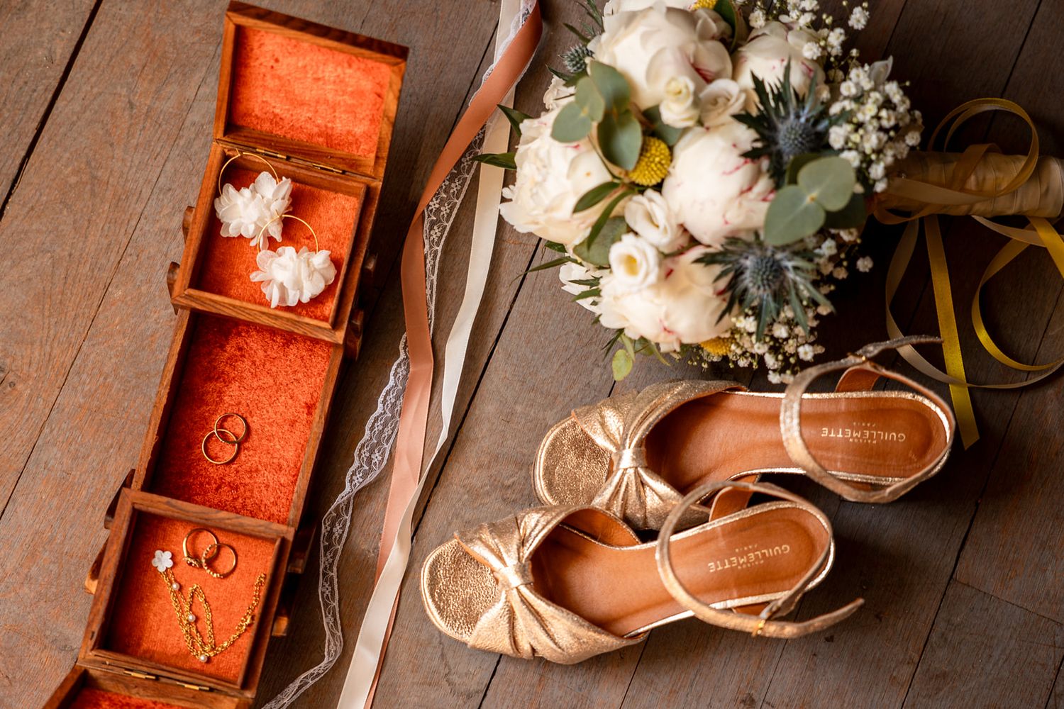 Wedding flat lay with gold heels, floral bouquet, jewelry box with rings, and ribbons on rustic wood floor.