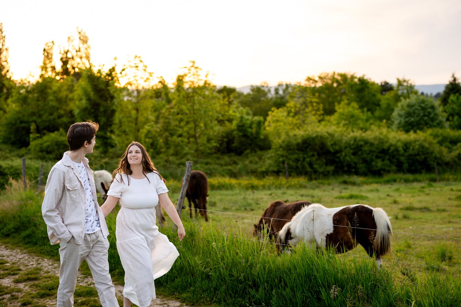Couple walking hand in hand near grazing horses at golden hour on a lush green farm.