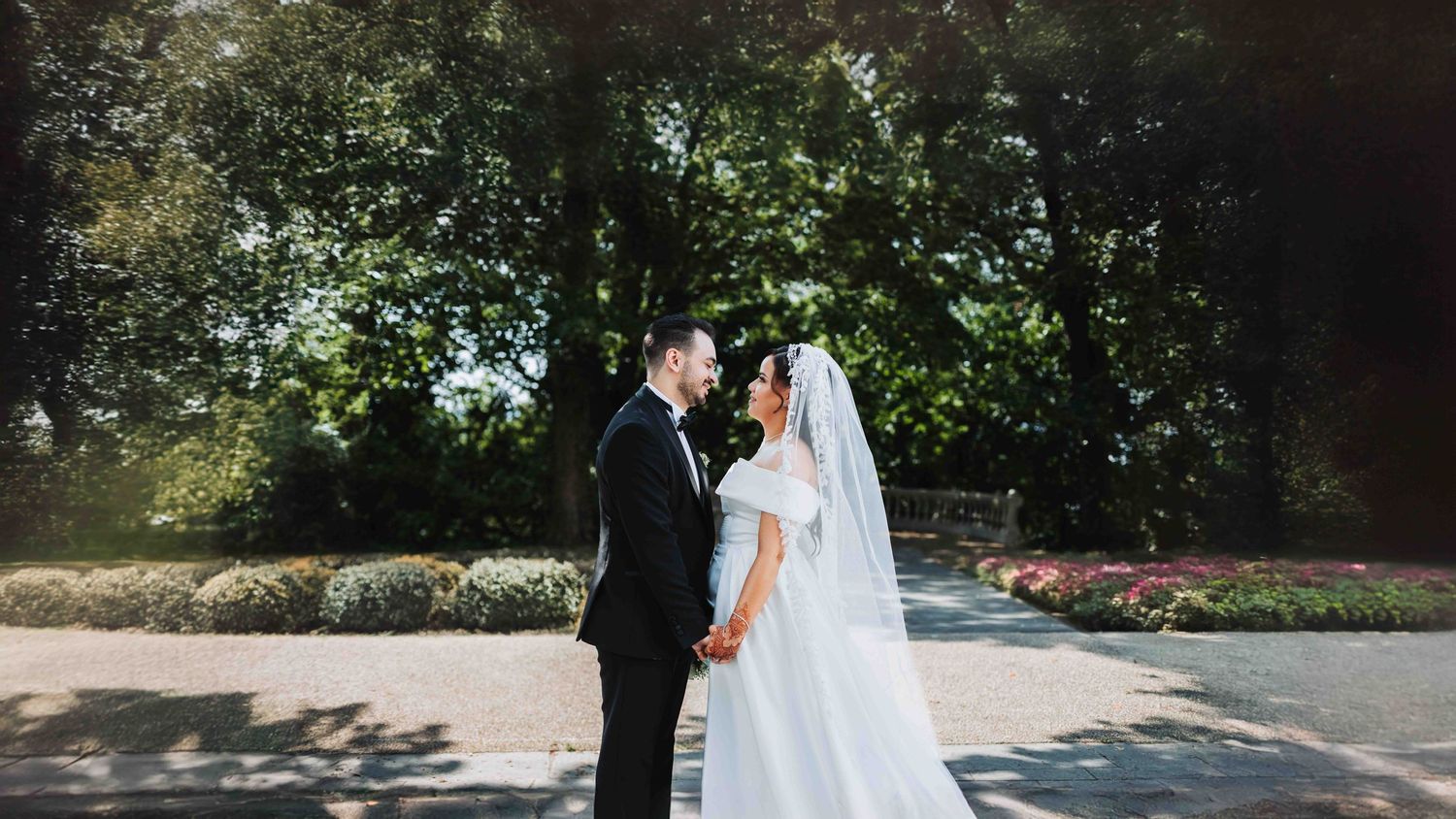 Wedding couple poses along tree-lined pathway in elegant outdoor setting.