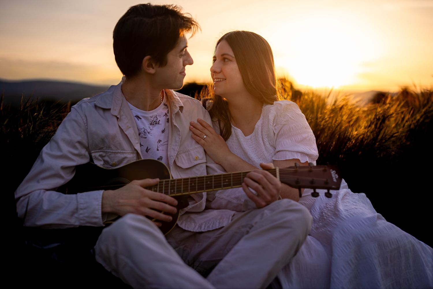 Couple sitting together at sunset, man playing acoustic guitar while woman listens, both wearing white in a romantic outdoor setting.