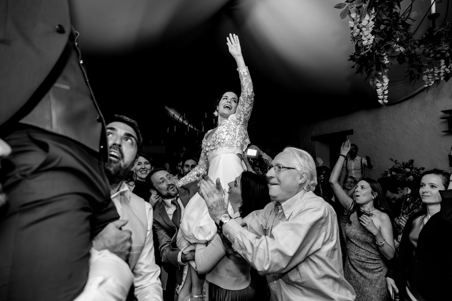 Black and white wedding photo of bride lifted by guests, arms raised joyfully, celebrating on the dance floor.