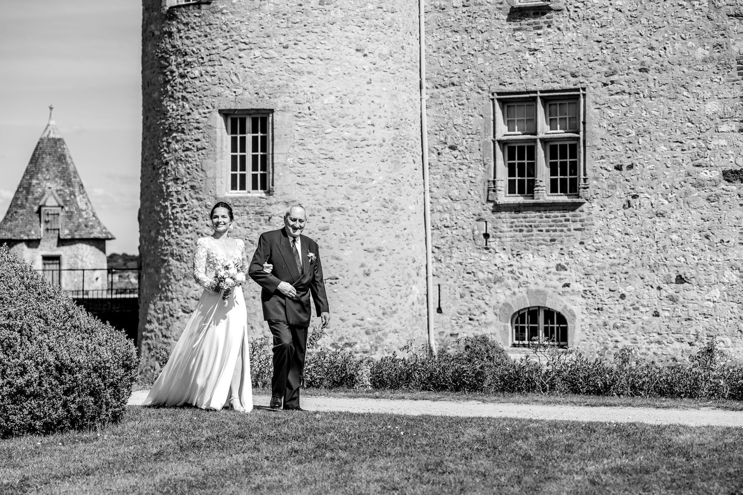 Black and white photo of a bride and her father walking toward a historic stone castle on a sunny wedding day.