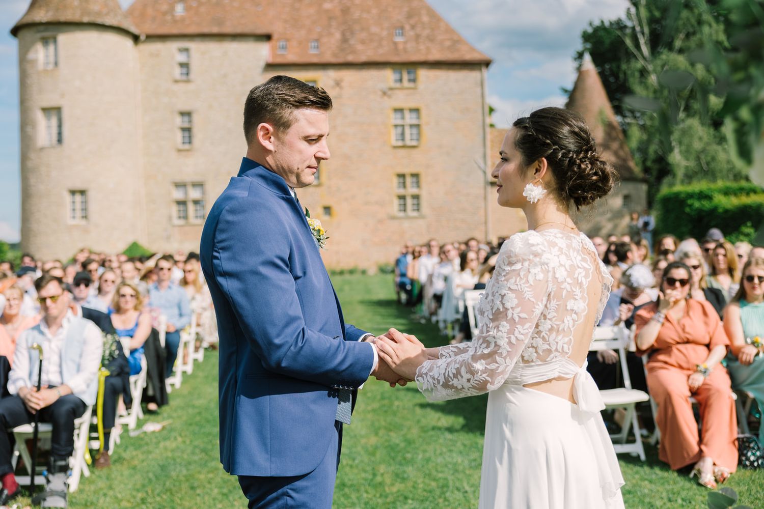 Couple exchanging vows at outdoor wedding ceremony with historic castle backdrop and seated guests.