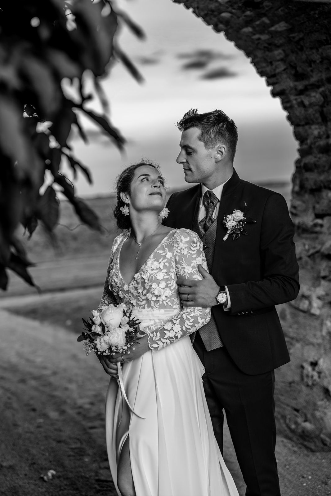 Black and white wedding photo of bride holding bouquet and groom in suit embracing under stone arch at dusk.