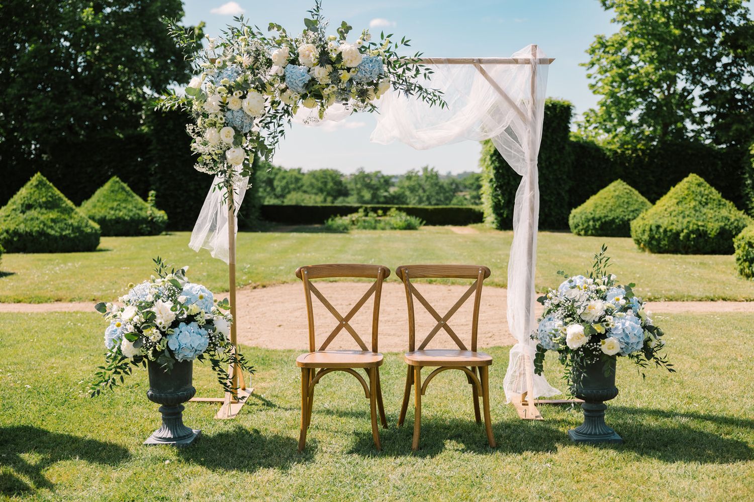 Elegant outdoor wedding ceremony setup with floral arch, two wooden chairs, and blue hydrangea arrangements on a lush green lawn.